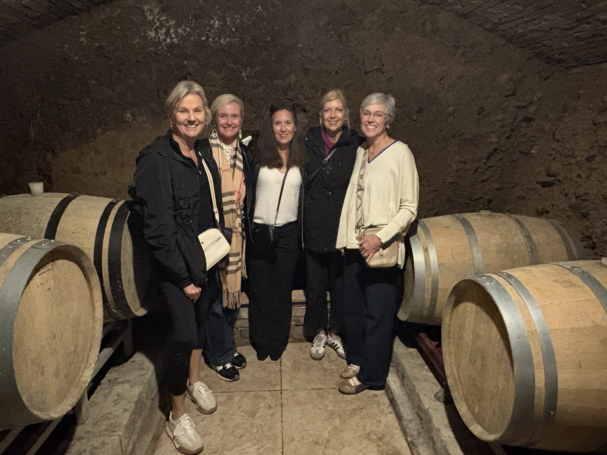Five women standing together inside a wine cellar with large wooden barrels in the background.