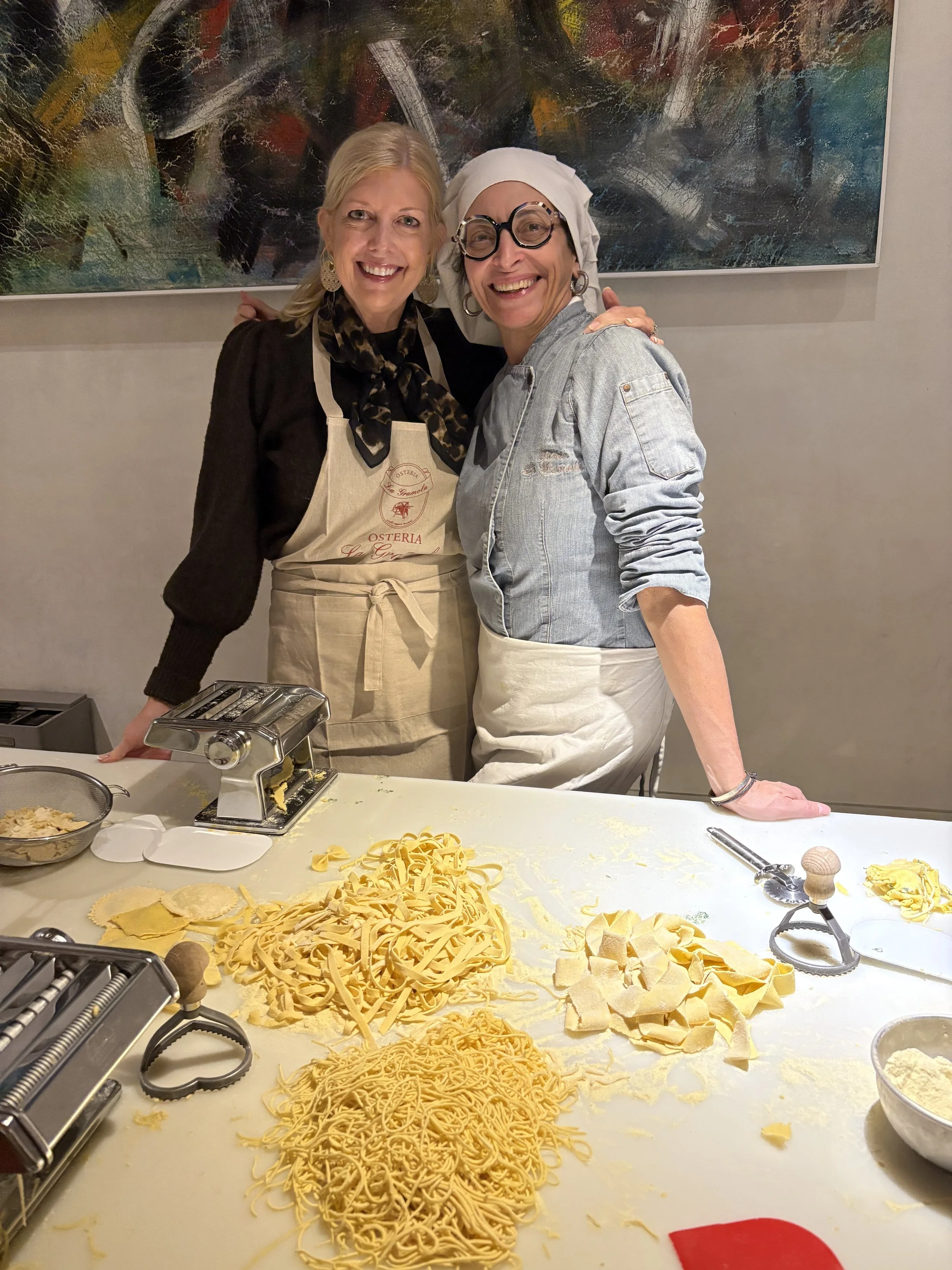 Two women in aprons smiling and posing behind a counter with pasta and cooking tools in a kitchen setting.