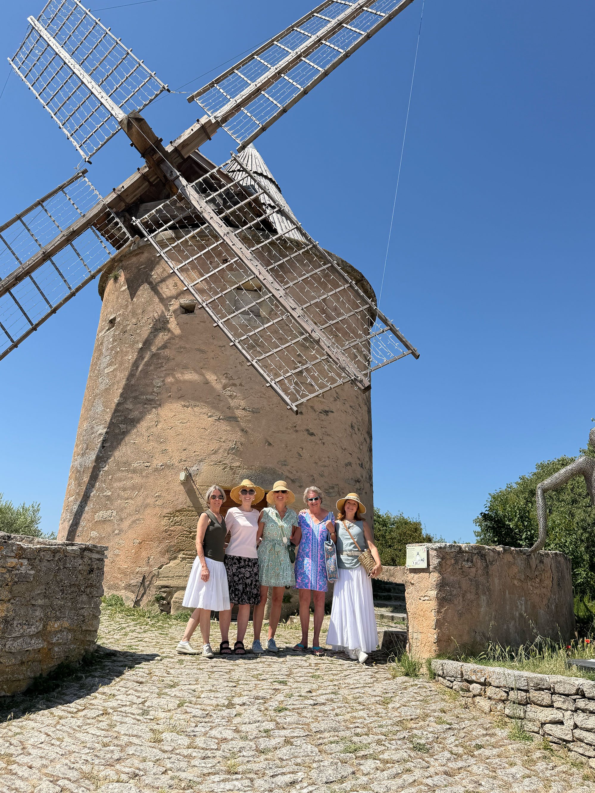 Group of five women standing in front of an old stone windmill with wooden blades, under a clear blue sky.