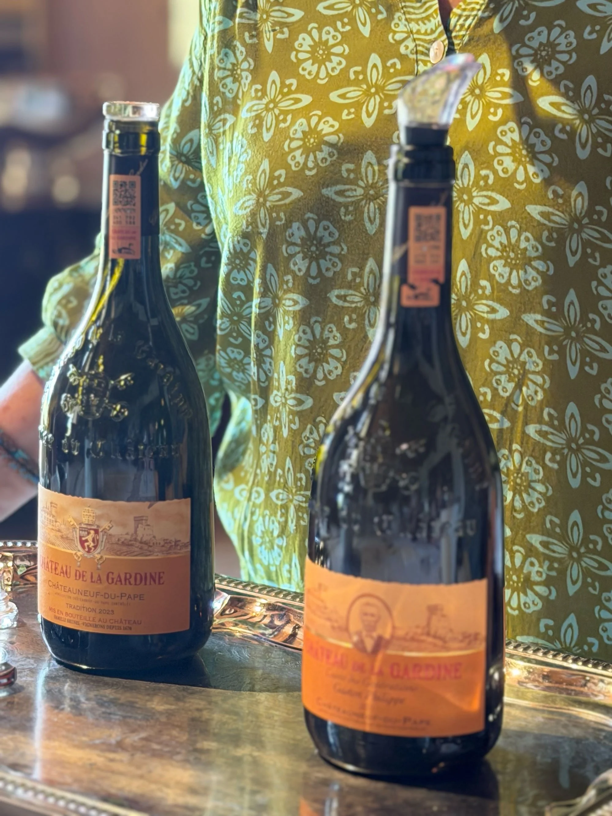 Two bottles of Château de la Gardine red wine on a table, with a person in a yellow shirt with a floral pattern in the background.