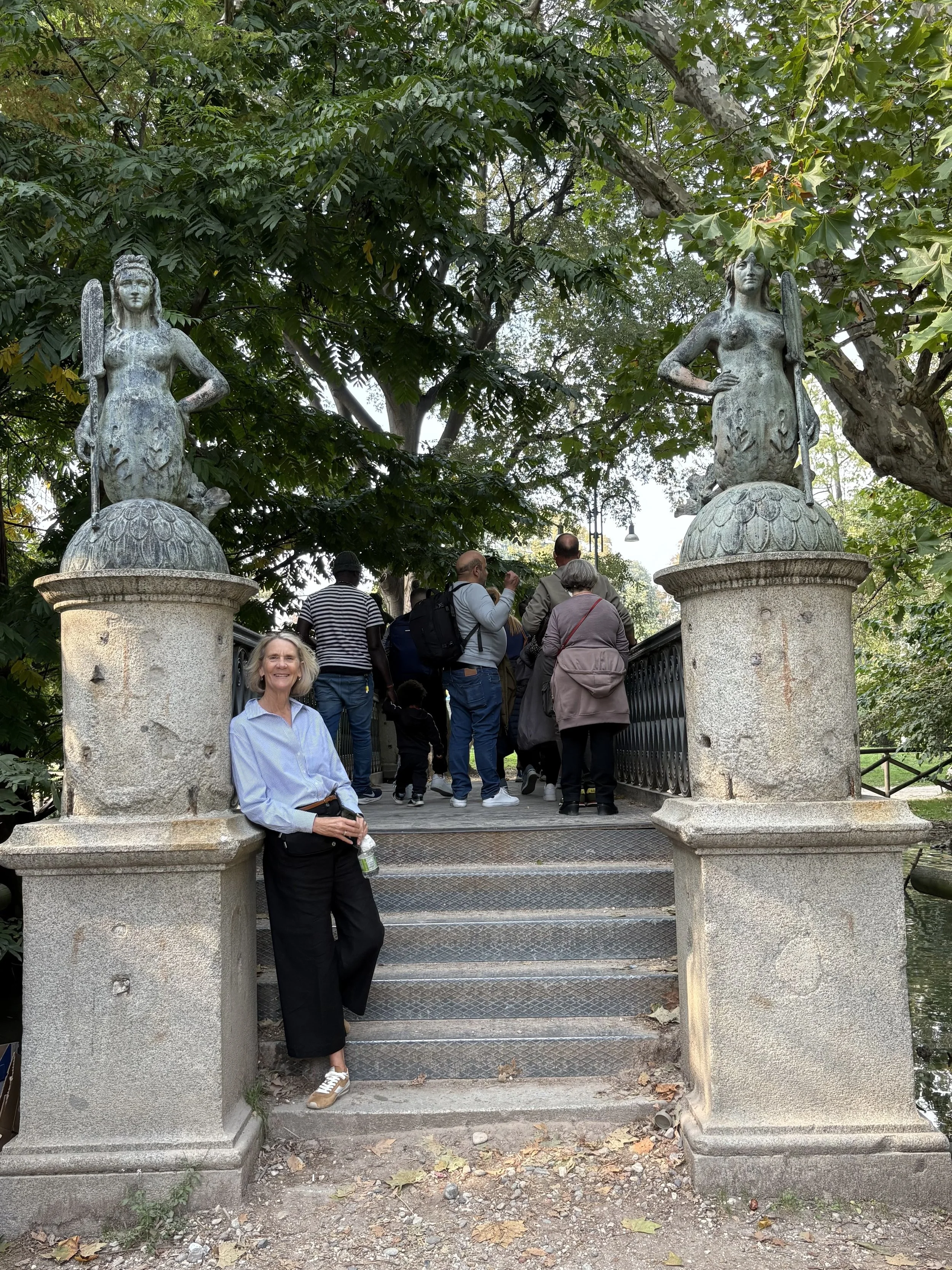 A woman stands at the bottom of a small stone staircase flanked by two statues of women holding spears. The statues are on large stone pedestals, surrounded by trees and foliage. A group of people is gathered on the bridge at the top of the stairs in