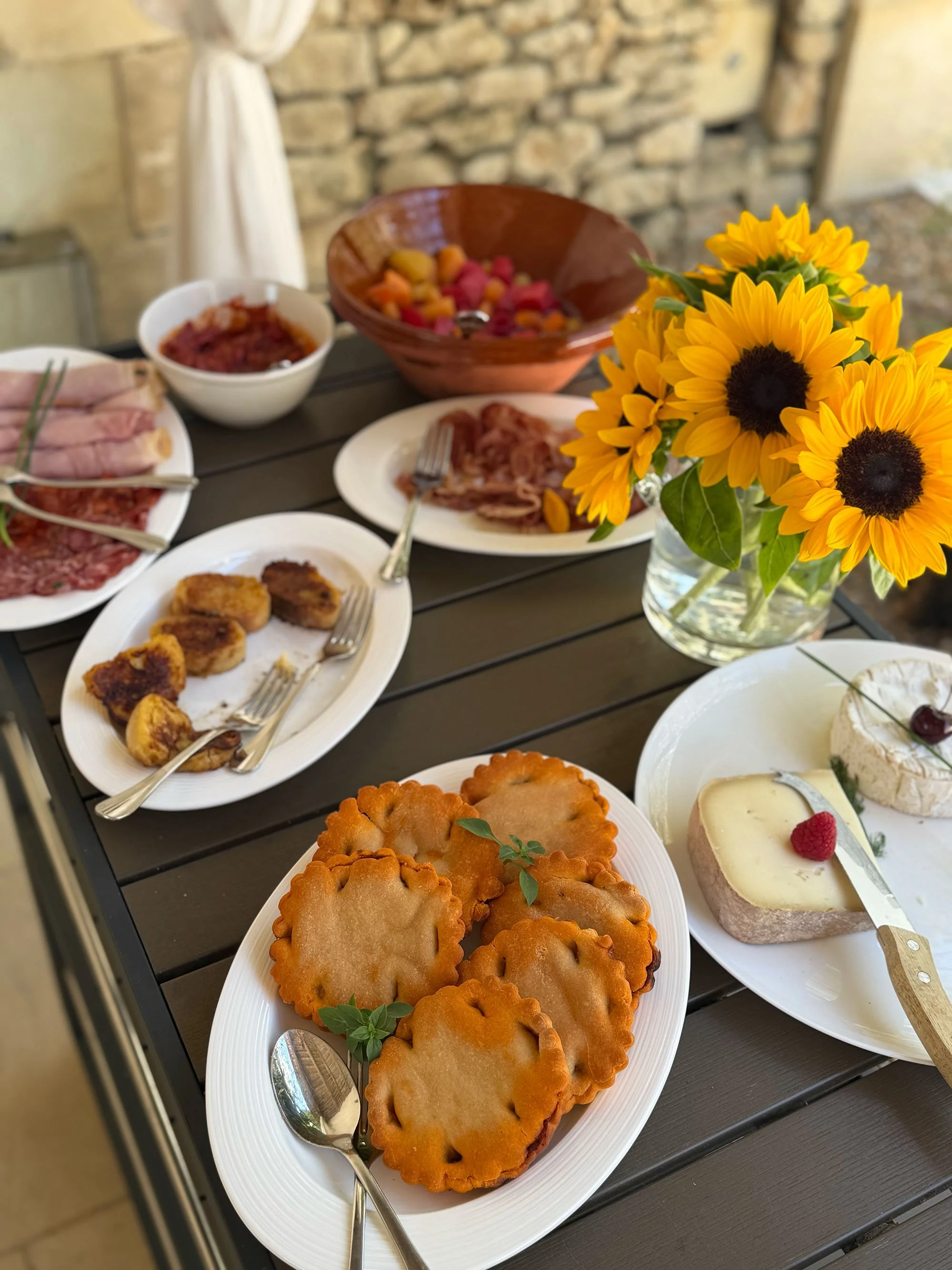A table of assorted foods including fried dough, cheese, cold cuts, a cheese wheel with a raspberry and a green leaf, and sunflowers in a vase.