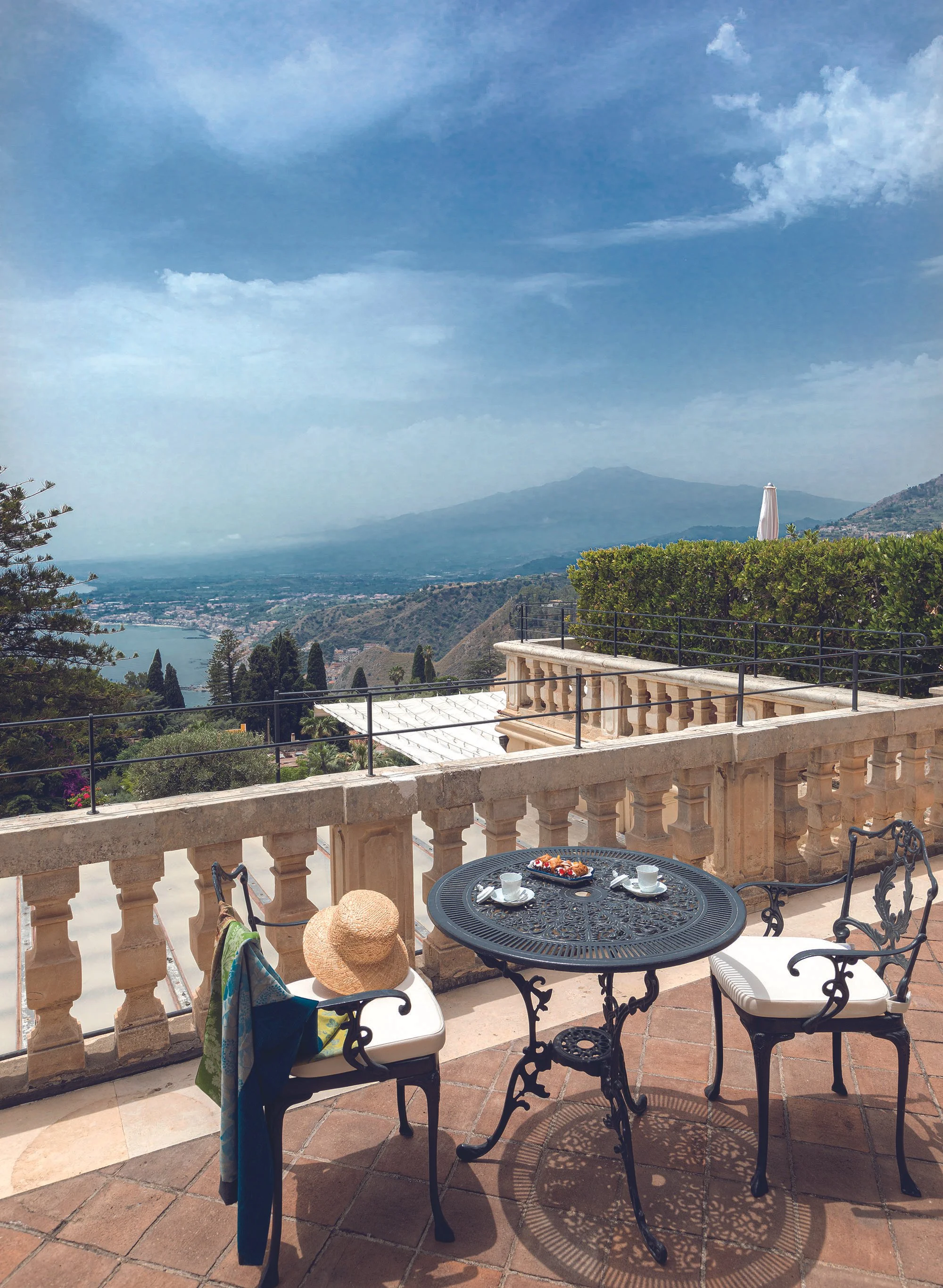 A balcony with a black wrought iron round table set for breakfast, surrounded by white and black chairs, with a scenic view of mountains, a lake, and a town below. There is a straw hat and a towel hanging on one chair, and a straw bag hanging on another. The table has a plate of pastries and cups of coffee.