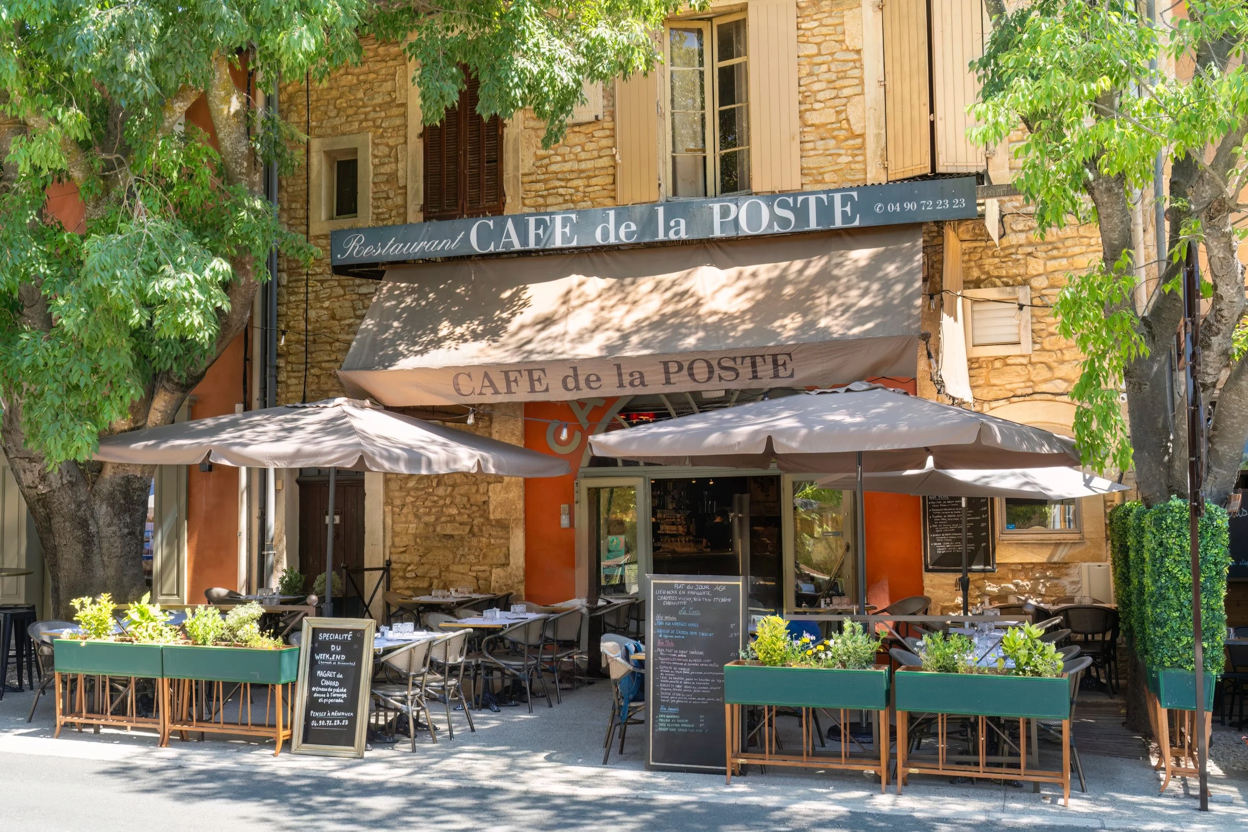 Exterior of a French cafe named 'Café de la Poste' with outdoor seating, umbrellas, and greenery in a sunny setting.