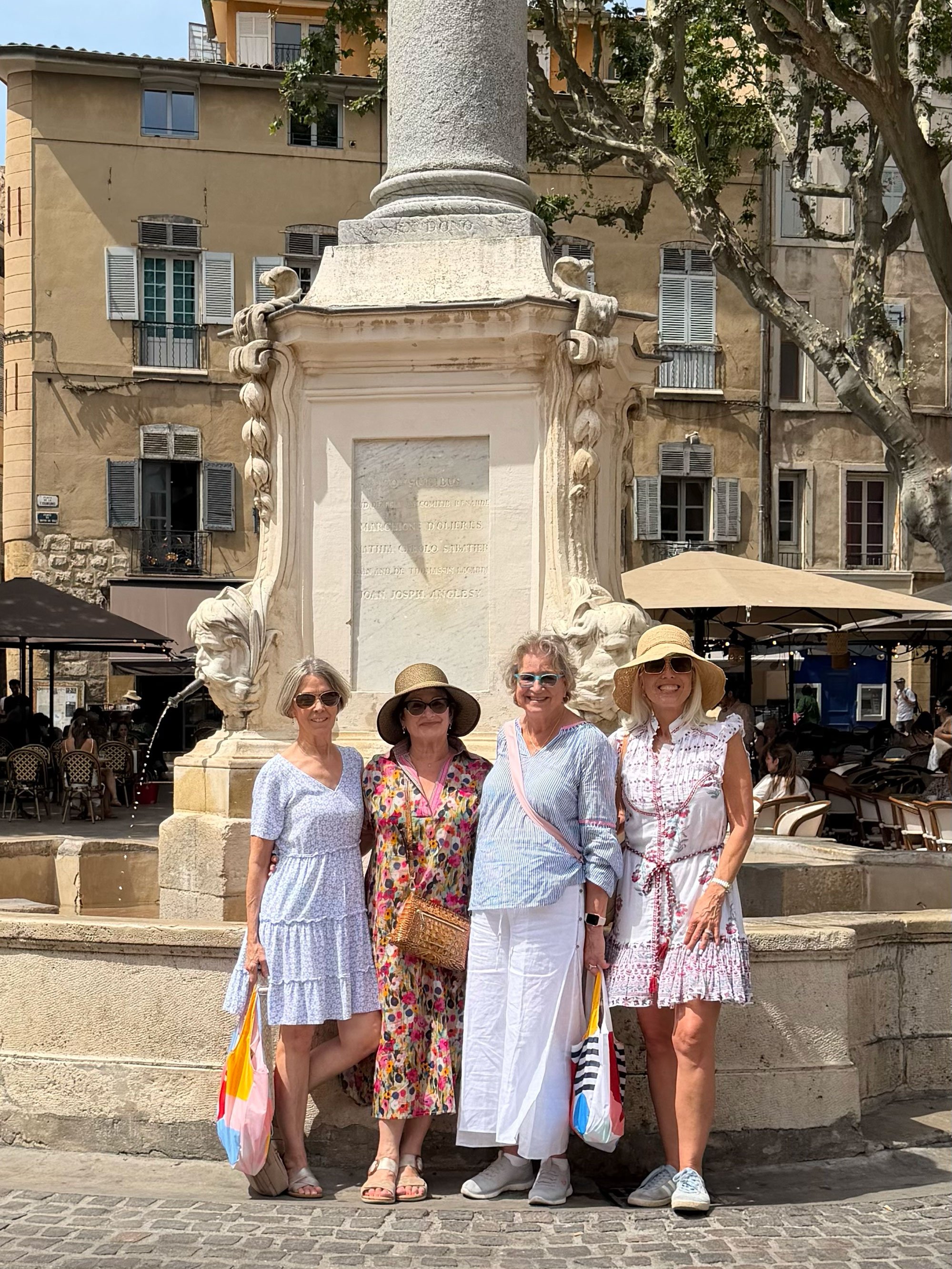 Four women standing in front of a historical stone monument in a city square, smiling and dressed in summer clothing and hats, with buildings and outdoor cafes in the background.