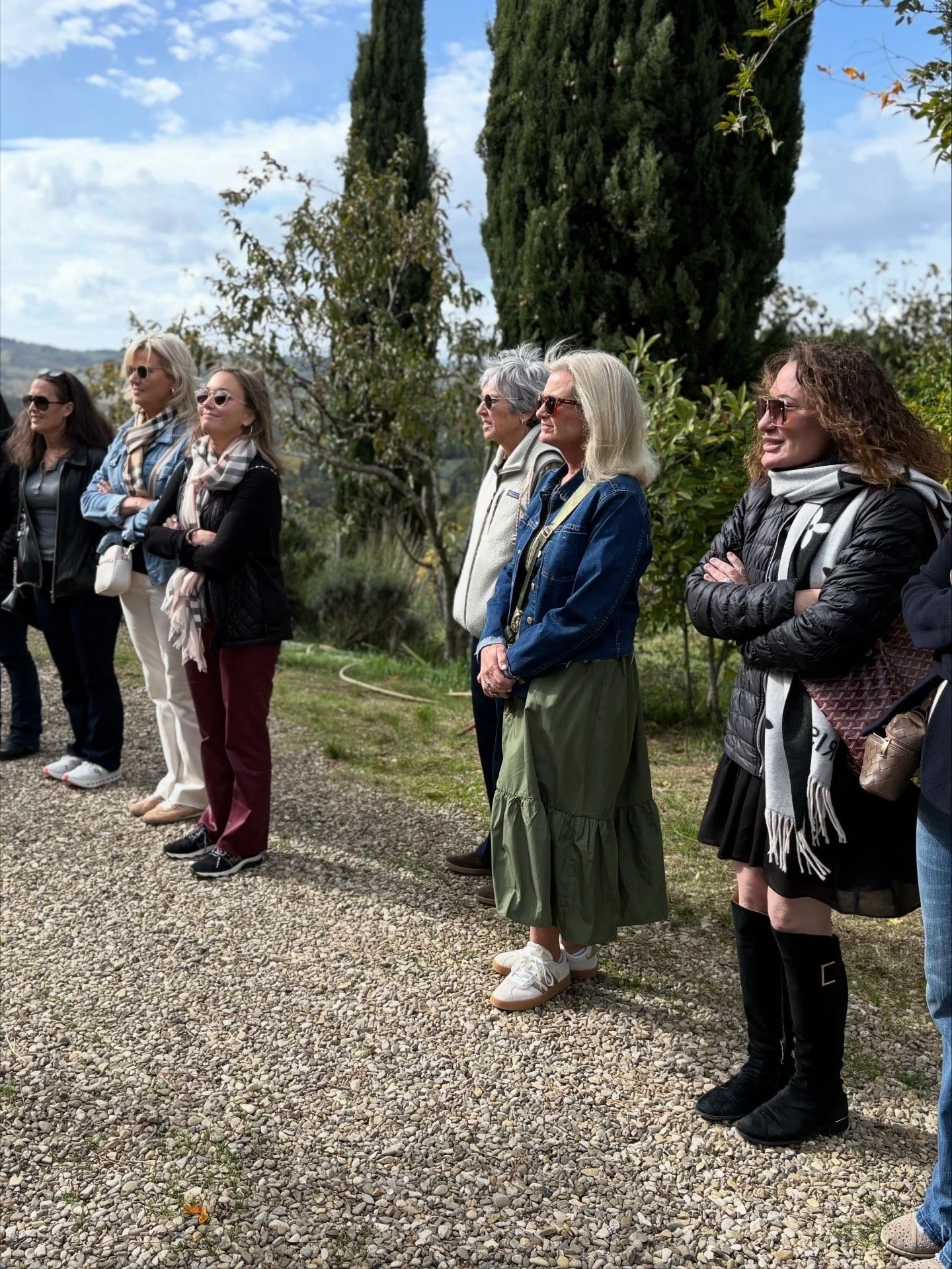 A group of women standing outdoors on a gravel path, facing to the right, with trees and bushes in the background under a partly cloudy sky.