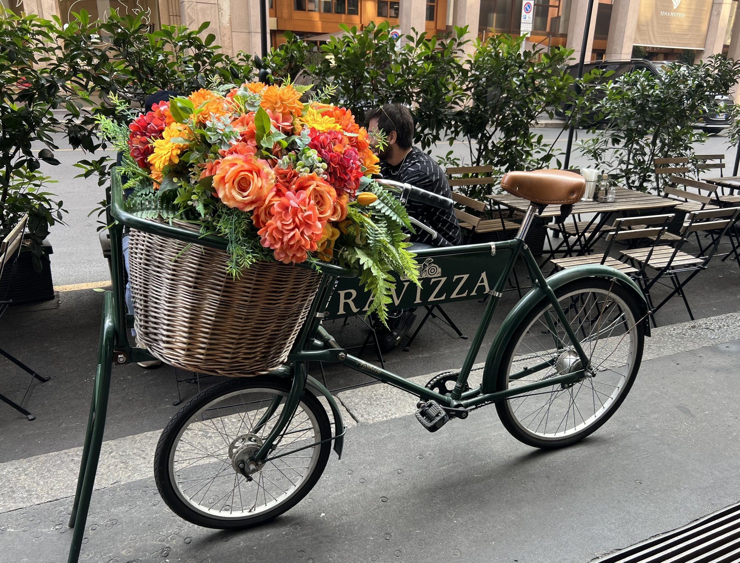 A green bicycle with a large basket of bright orange, yellow, pink, and red flowers parked outside a restaurant with outdoor seating. There are two men seated at a table behind the bike, one wearing a face mask.