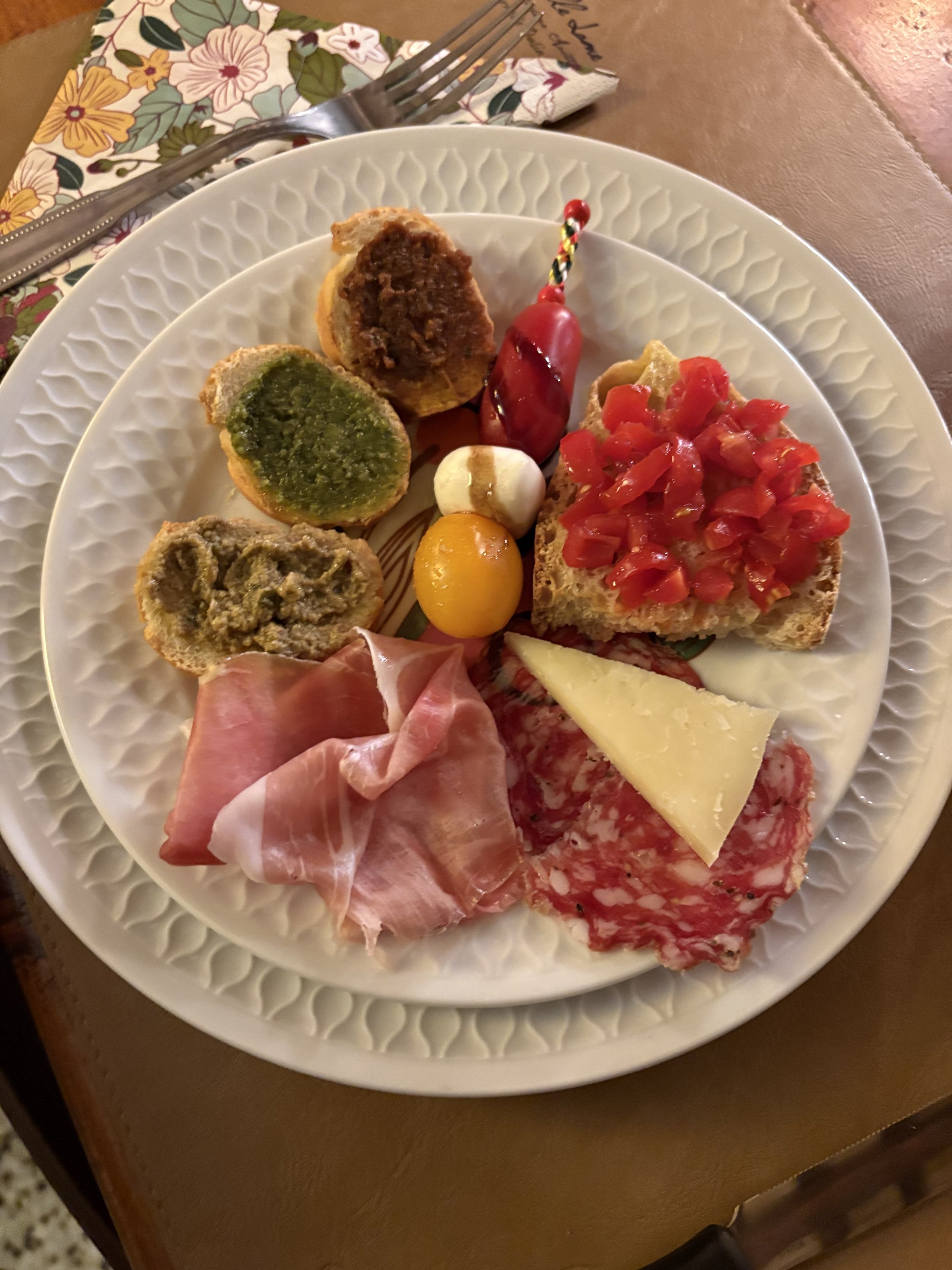 A white plate with assorted Italian antipasto including cured meats, cheeses, bread topped with diced tomatoes, roasted eggplant, green pesto, and a small skewer of cherry tomatoes and mozzarella balls.