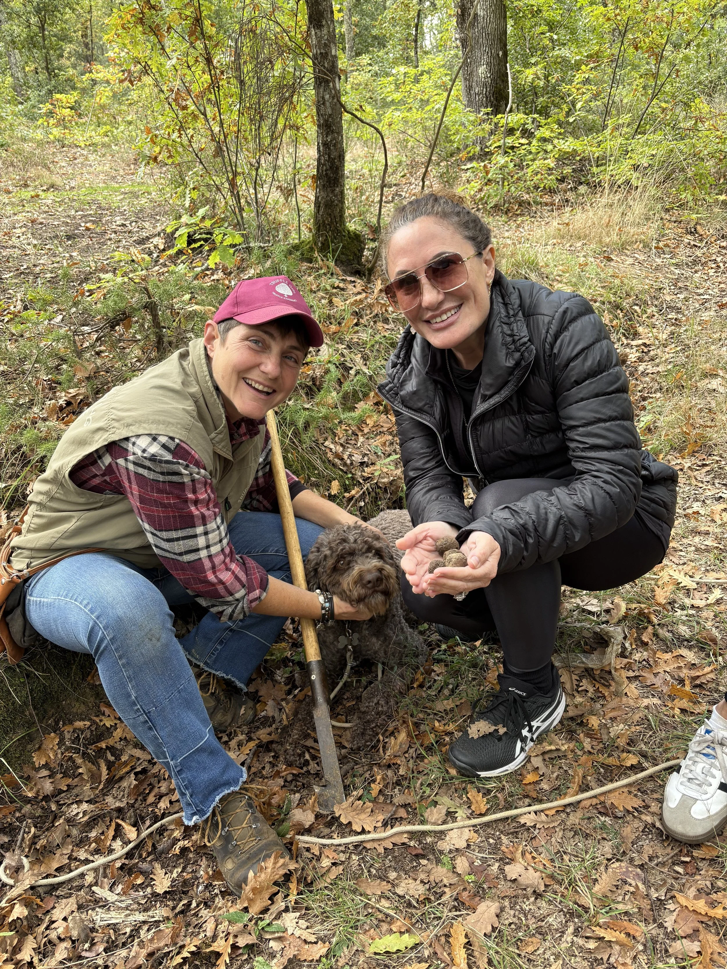 Two women and a dog in a forest collecting small objects or rocks; one woman is holding a dog and the other showing the objects in her hand.