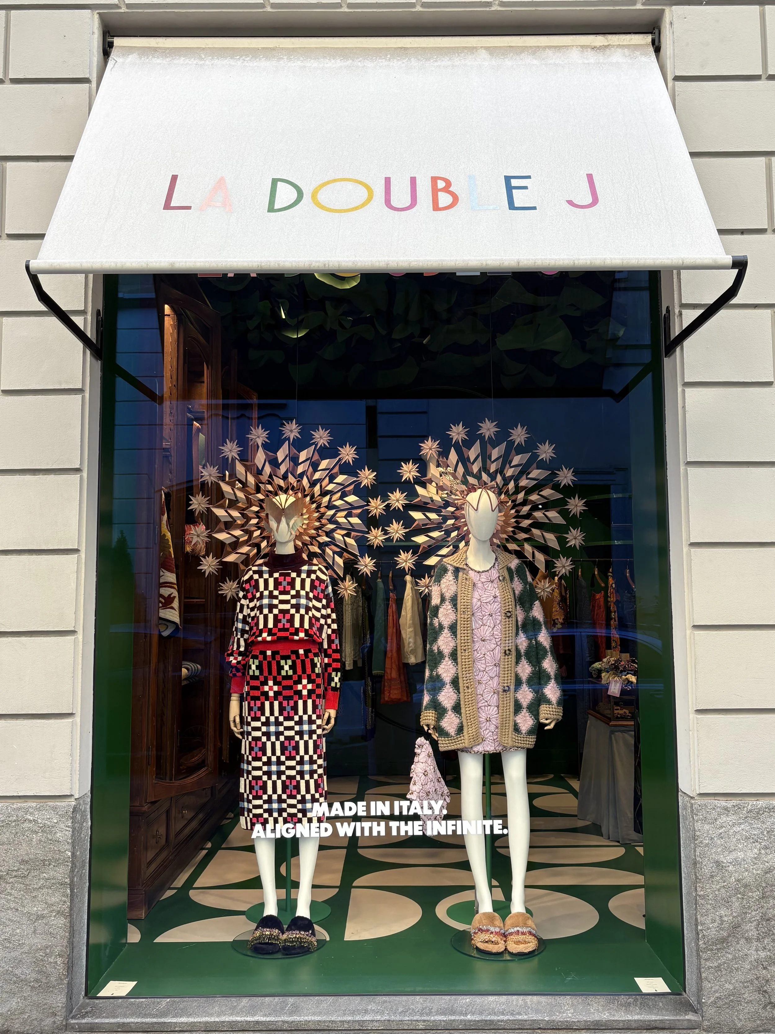 Store window display of two mannequins dressed in high fashion outfits with the sign 'LA DOUBLE J' on the awning. The mannequins have artistic headpieces resembling starbursts.