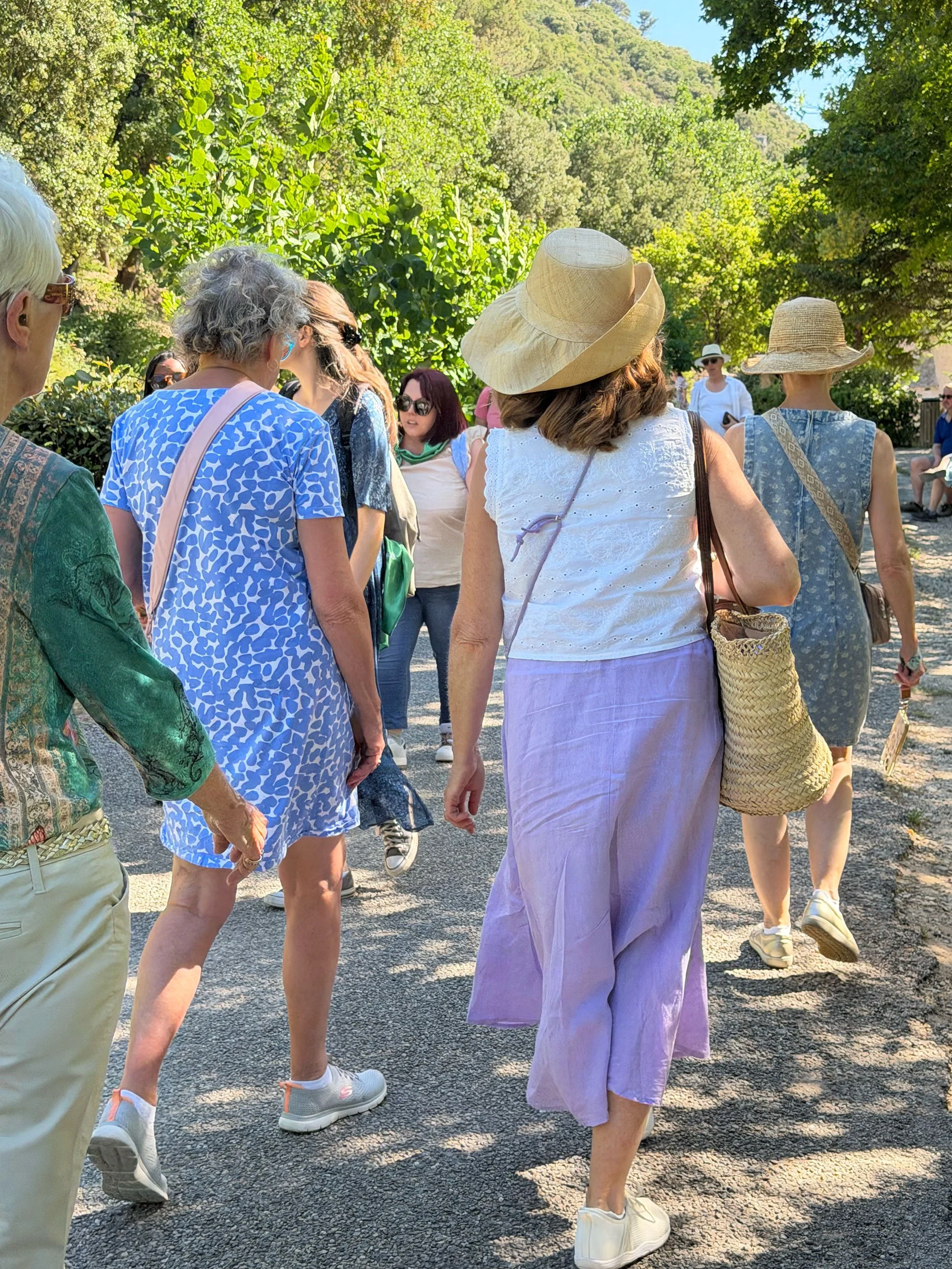A group of people, mostly women, gathered outdoors in a sunny setting with lush green trees in the background. Many are wearing hats and sunglasses, and they appear to be engaged in conversation or listening to someone. The scene suggests a casual so