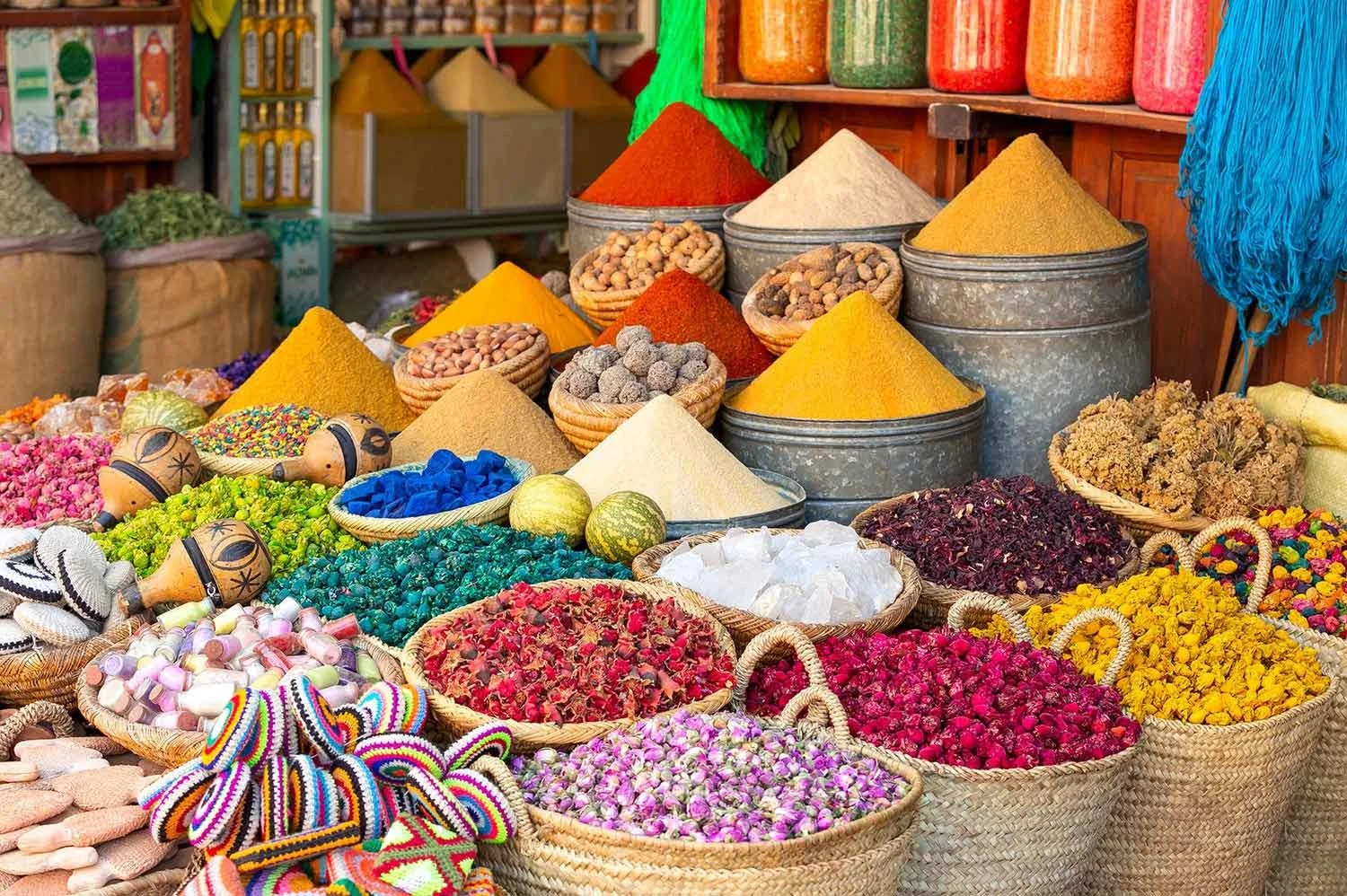 Colorful spices in metal and wicker containers at a marketplace, including red, yellow, white, and green powders, and dried herbs or flowers in Chefchaouen, Fez, Marrakesh.