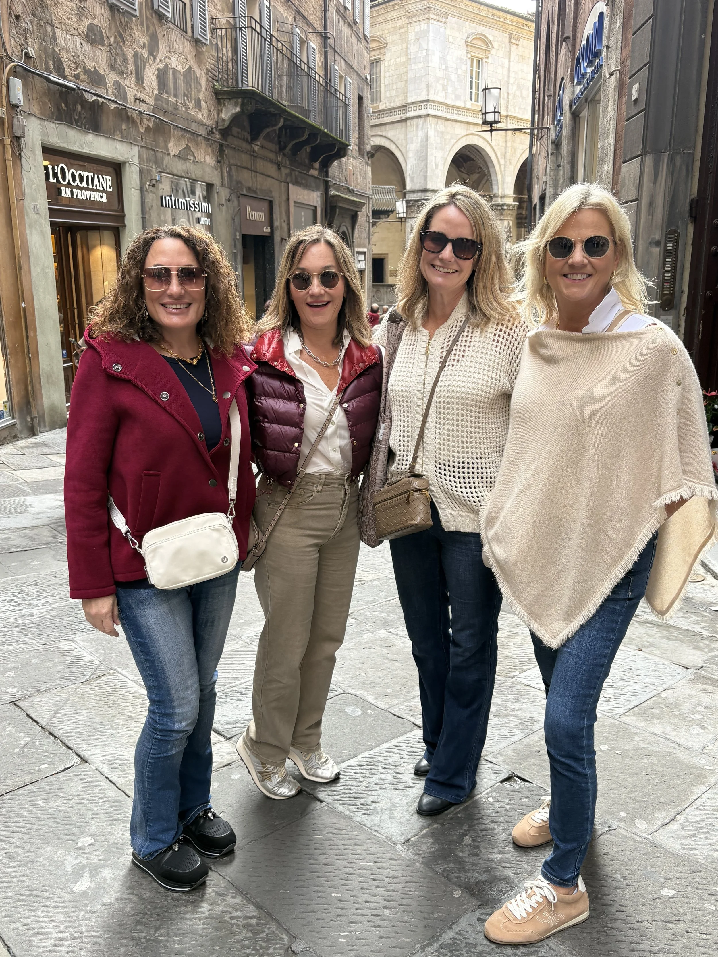 Four women standing on a cobblestone street, smiling at the camera, with storefronts and old buildings in the background.