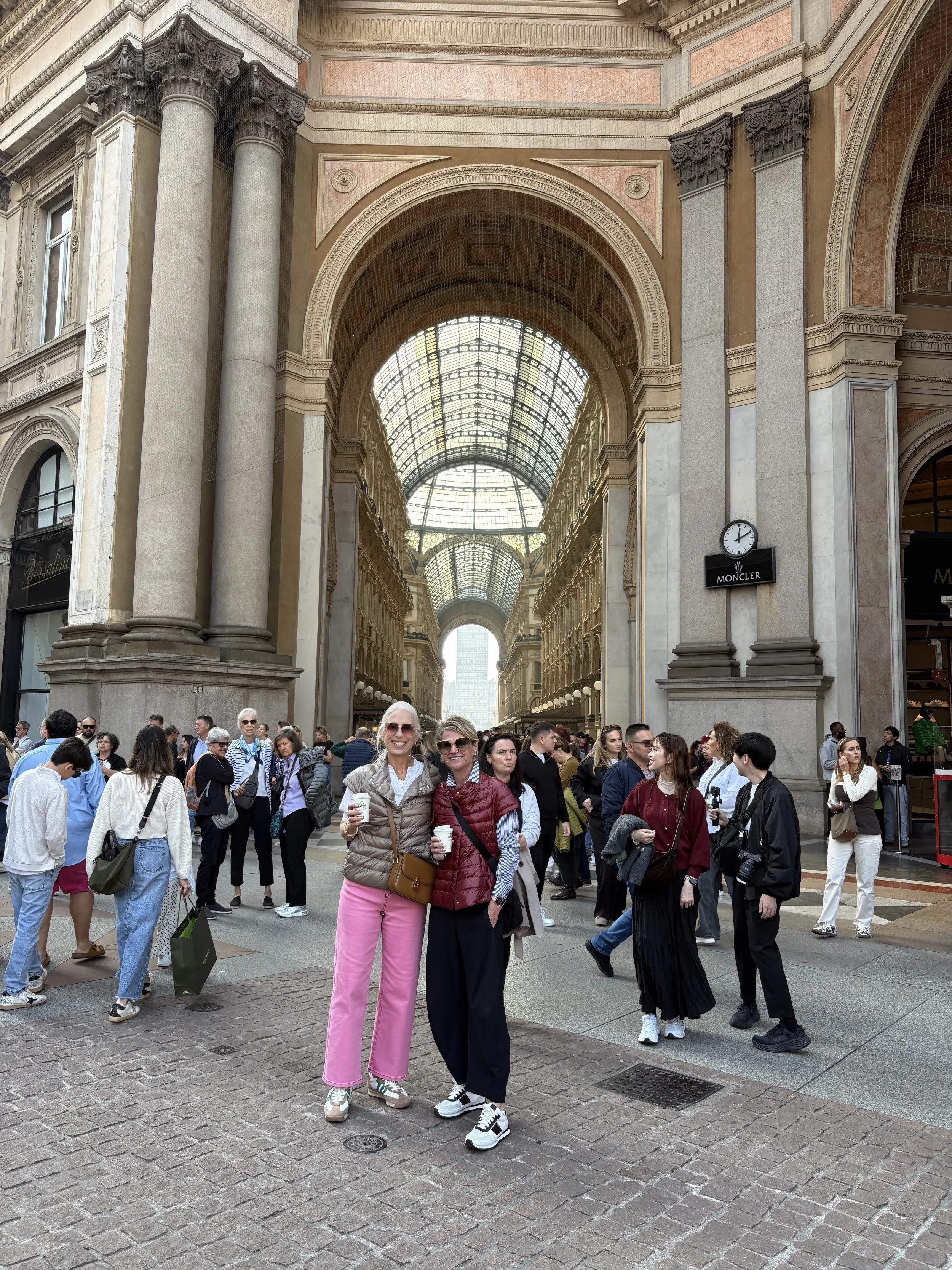 Two women smiling and holding coffee cups in front of a busy shopping arcade with a glass roof, columns, and an ornate ceiling.