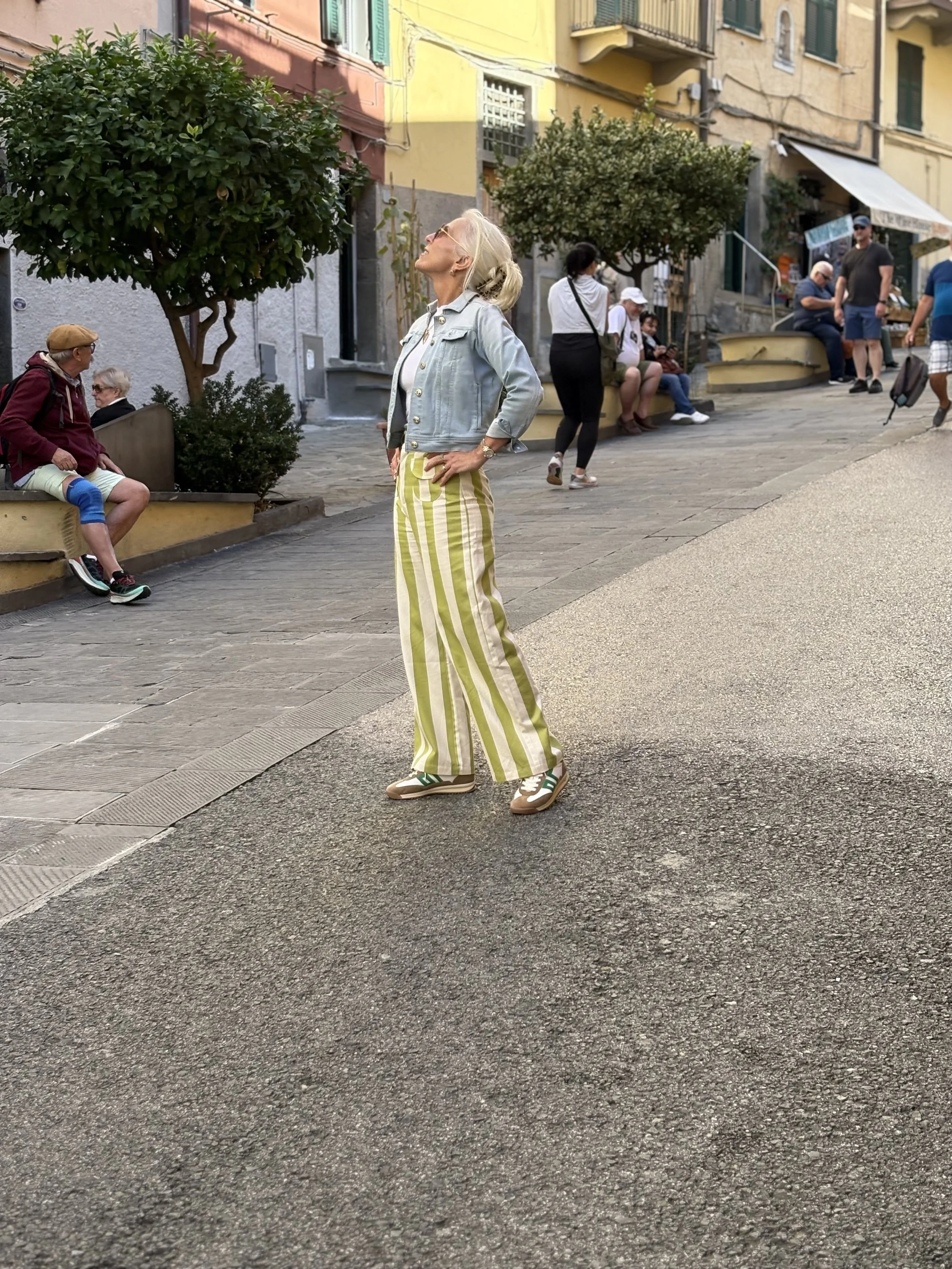 A woman with gray hair stands on a street with her hands on her hips, looking up. She is wearing a light denim jacket, yellow and white striped wide-leg pants, and sneakers. Several people are sitting and walking in the background, with colorful buil
