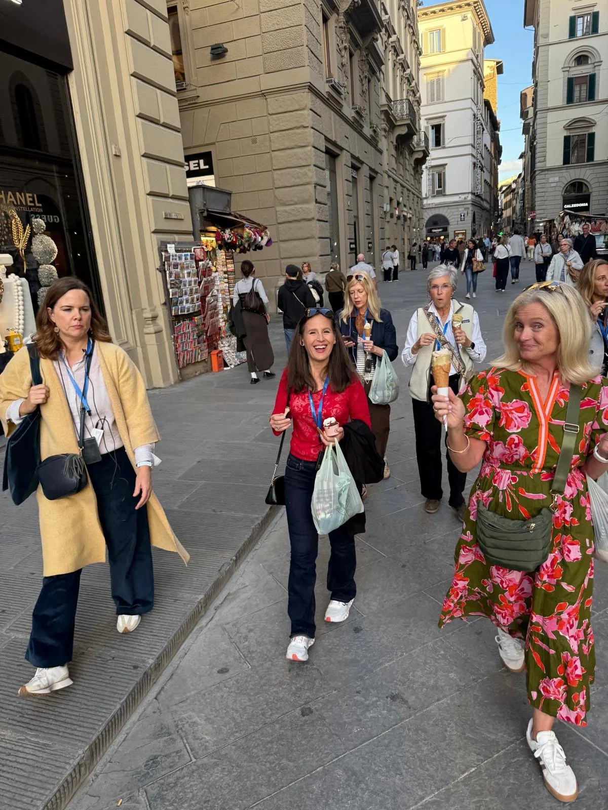 Group of women walking down a city street, some holding ice cream, with historical buildings and shops in the background.