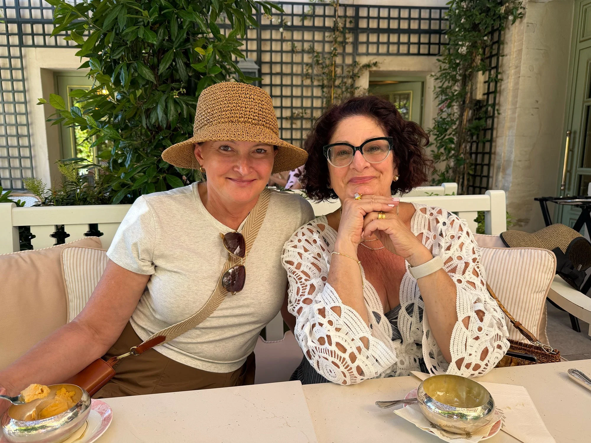 Two women sitting closely at a table outdoors, smiling, with bowls of food in front of them, surrounded by greenery and a lattice wall.
