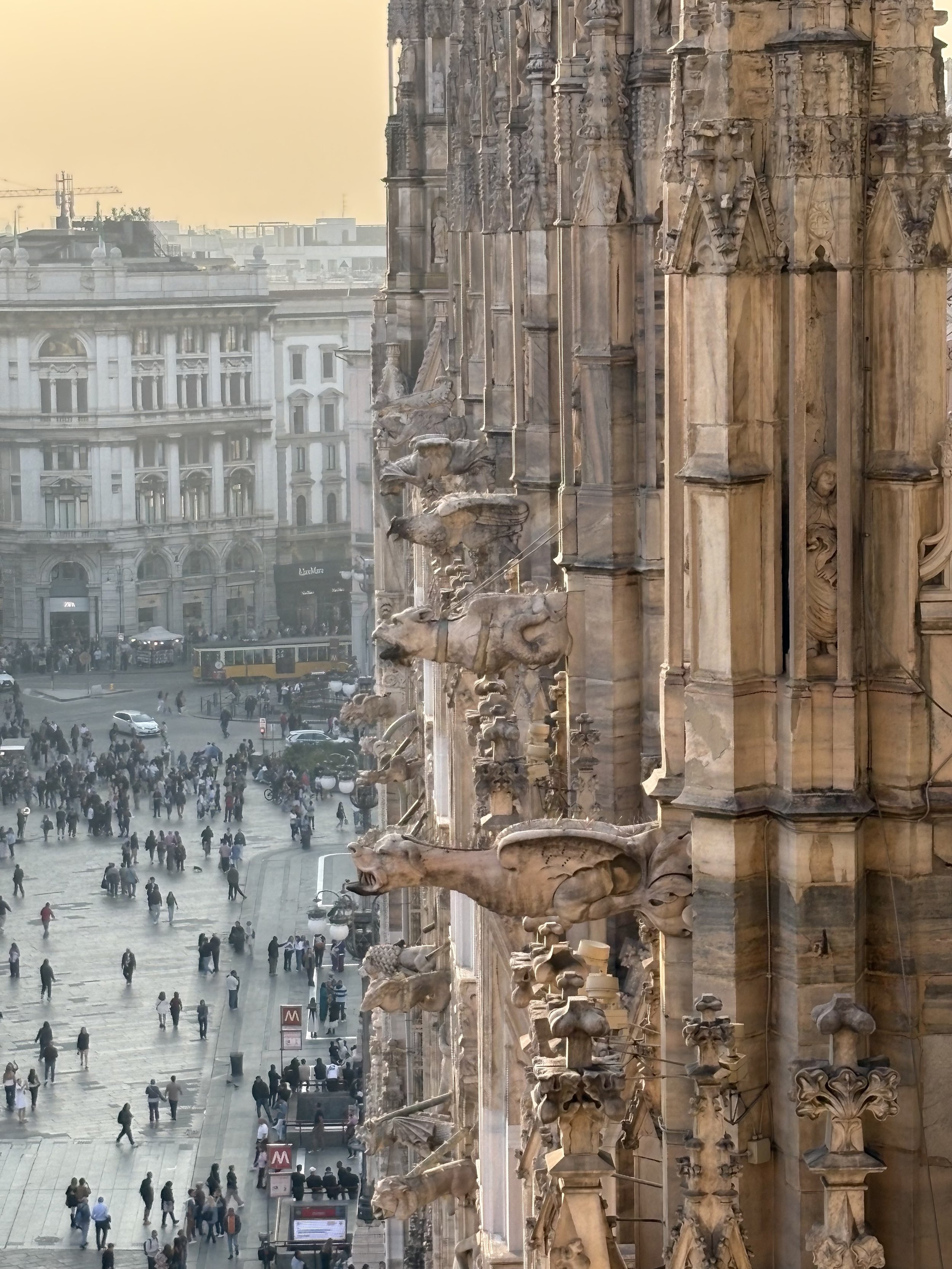 Close-up view of the Gothic-style architecture of the Milan Cathedral (Duomo di Milano), with detailed stone sculptures of animals and mythical creatures on the facade and a busy city square with people below.