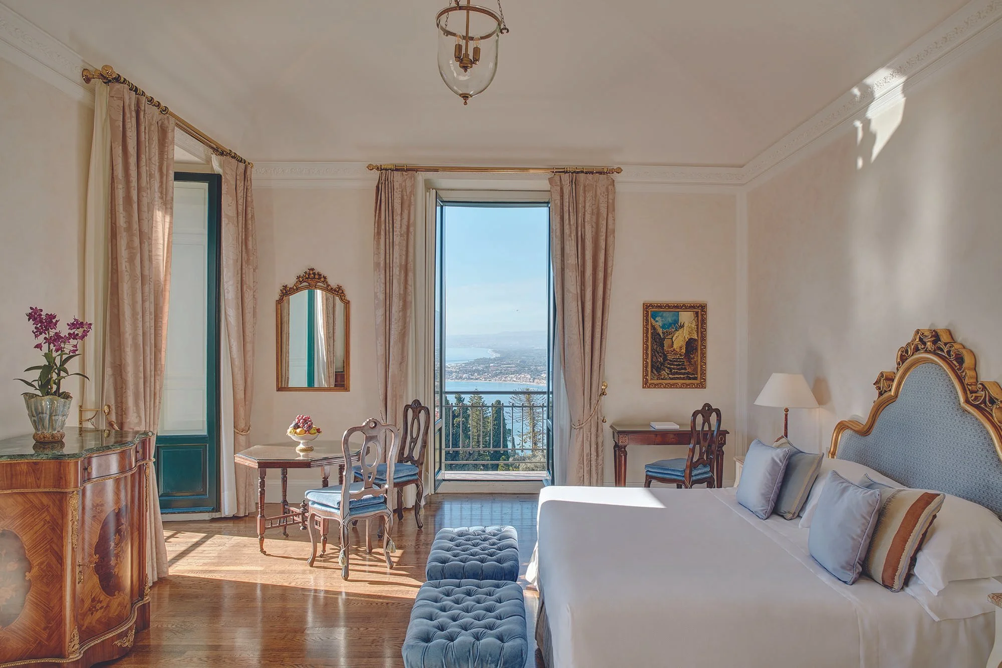 Elegant bedroom with a large window showing a scenic view of water and land, decorated with pink curtains, a vintage mirror, a wooden desk with a bowl of fruit, a blue upholstered bed and chairs, and a chandelier.