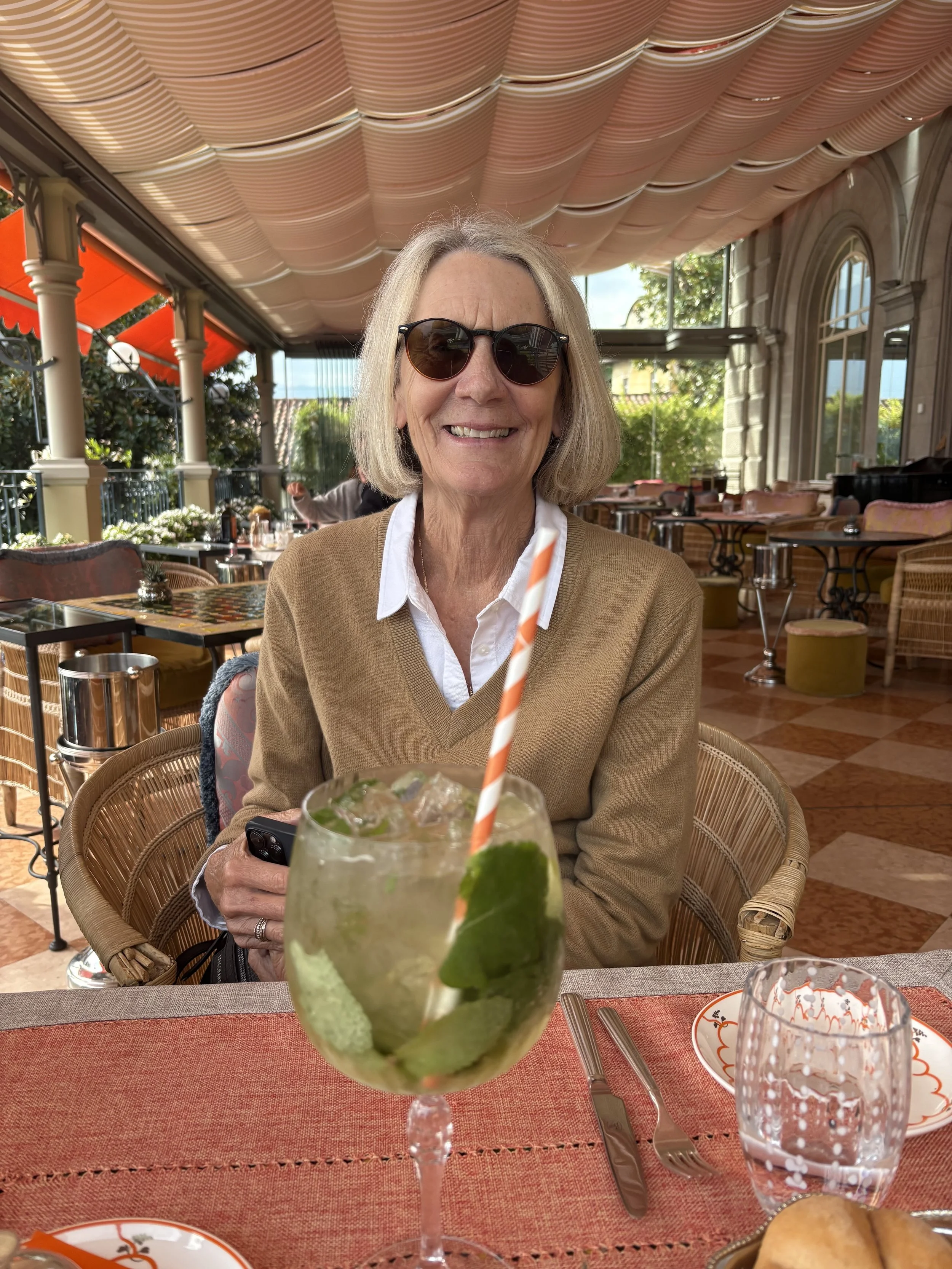 An elderly woman wearing sunglasses and a beige sweater, smiling while sitting at a restaurant table. There is a cocktail with lime and mint in the foreground, and the restaurant has a decorated ceiling, large windows, and outdoor views.