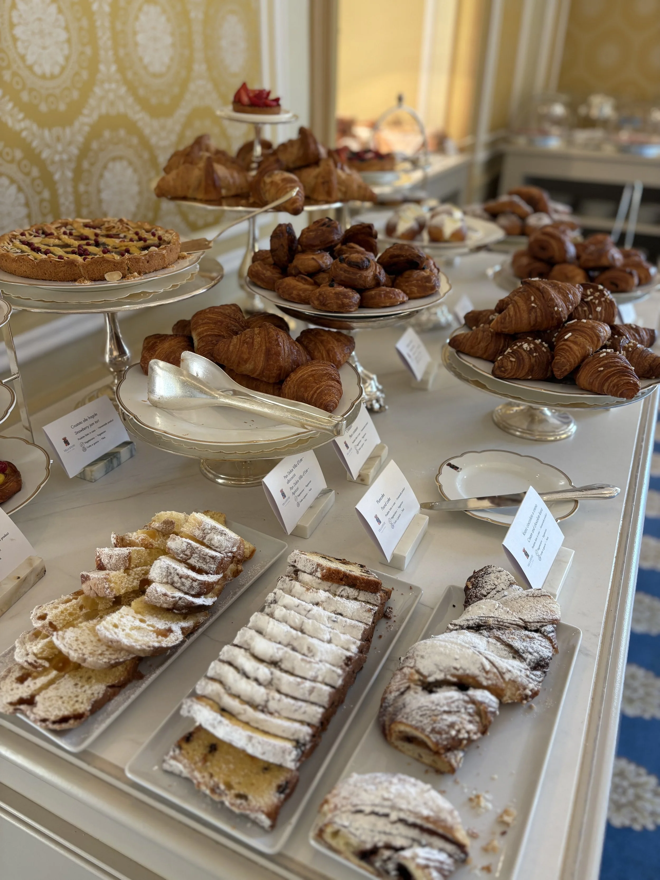 Assorted pastries including croissants, Danish pastries, and fruit tarts displayed on elegant cake stands and plates at a dessert buffet table.