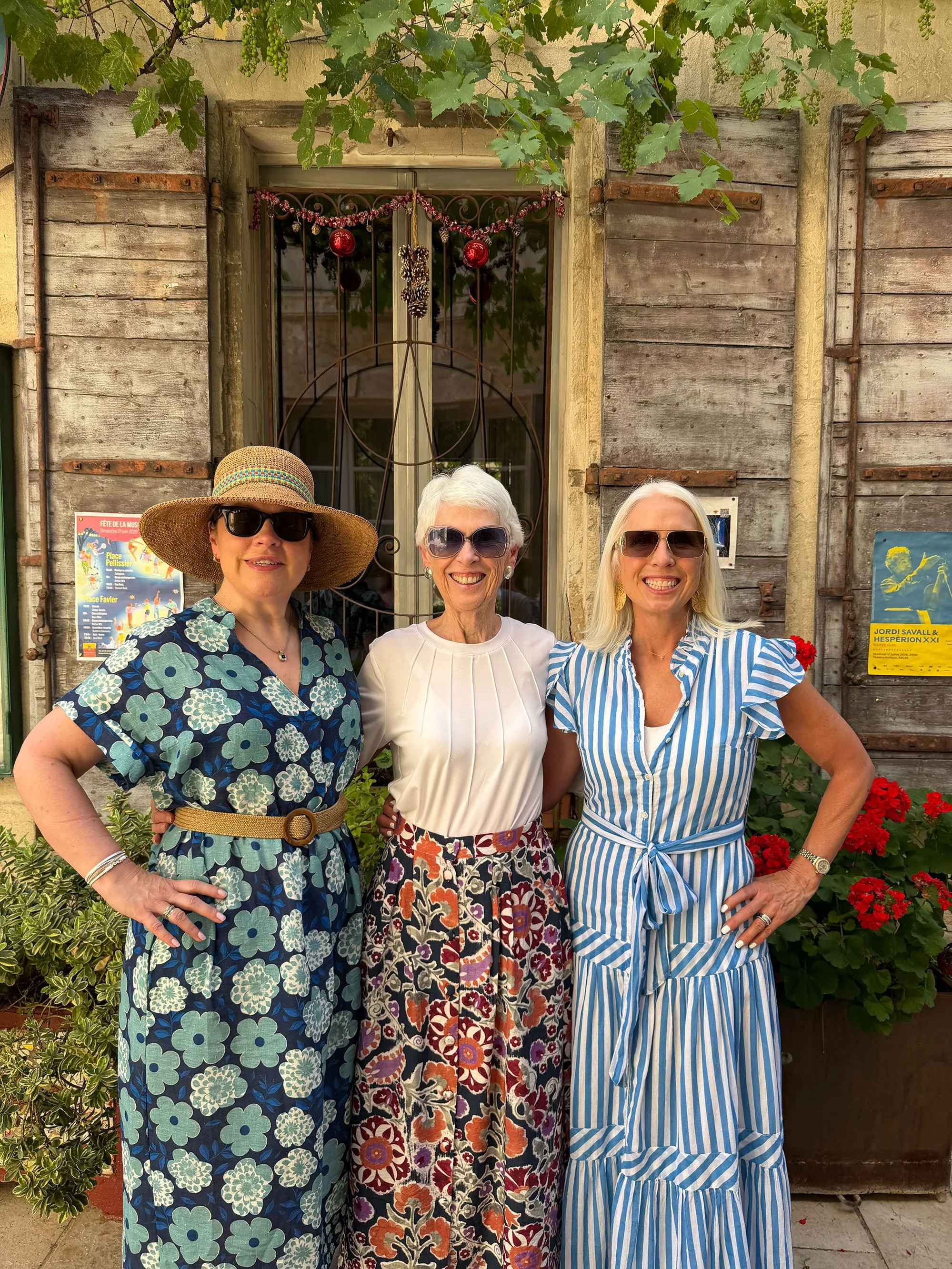 Three women smiling and posing together outdoors, standing in front of a rustic wooden wall with greenery and flowers, wearing colorful summer dresses and sunglasses.