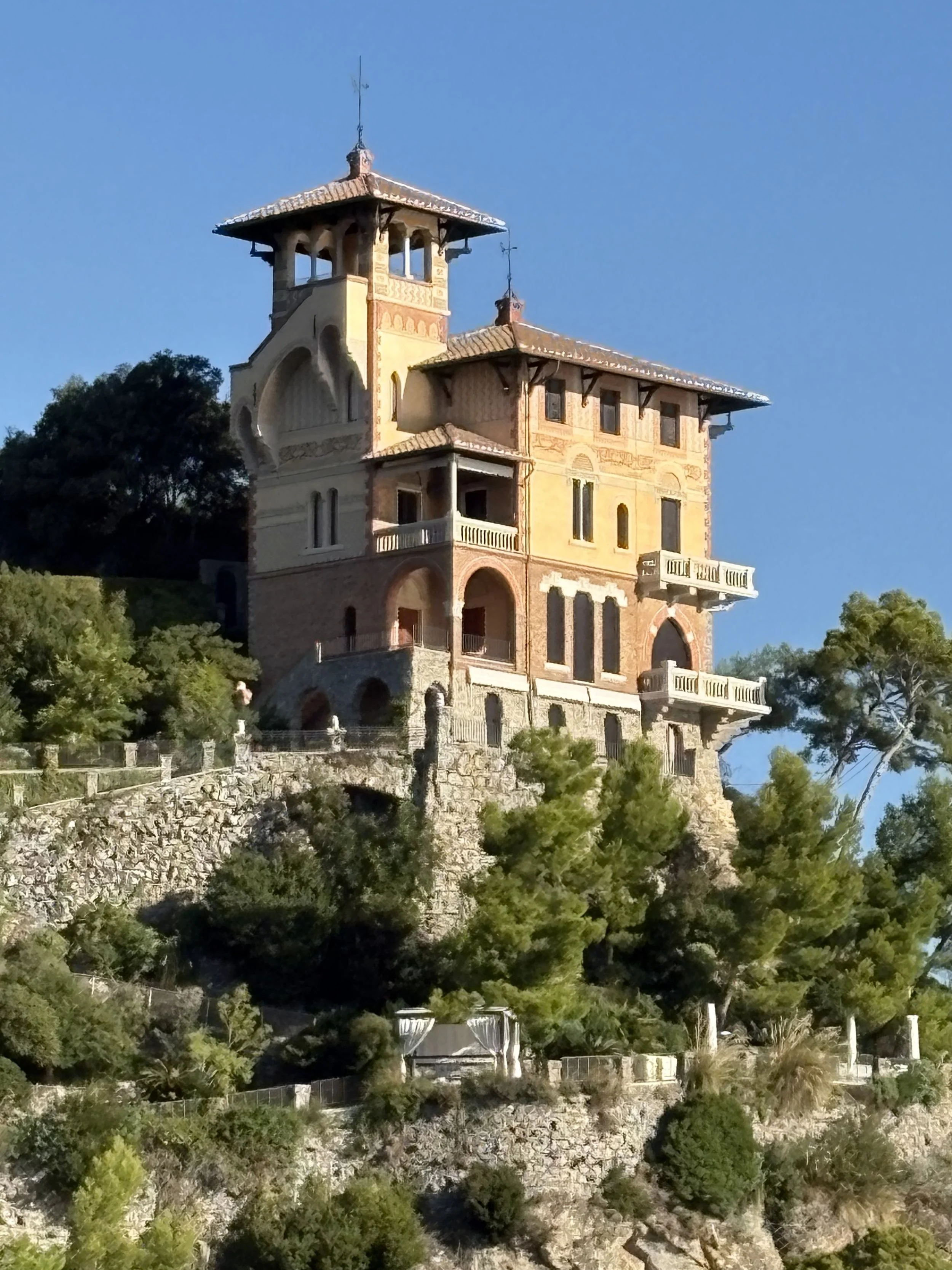 A large, ornate house built on a hill with trees and shrubs surrounding it, featuring balconies, multiple windows, and a tower with a weather vane on top, against a clear blue sky.