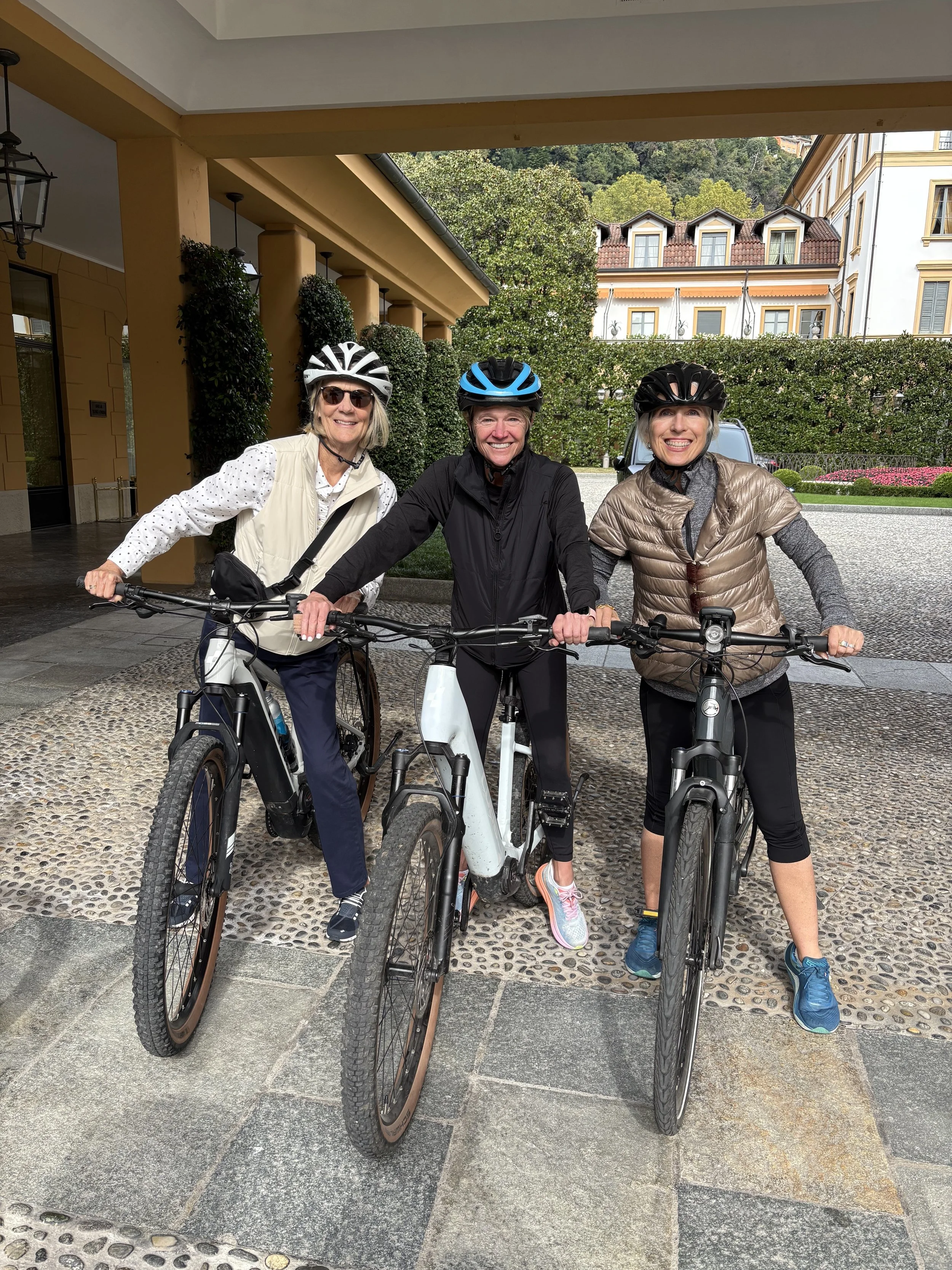 Three women with bikes smiling outdoors in front of a building with greenery and trees.