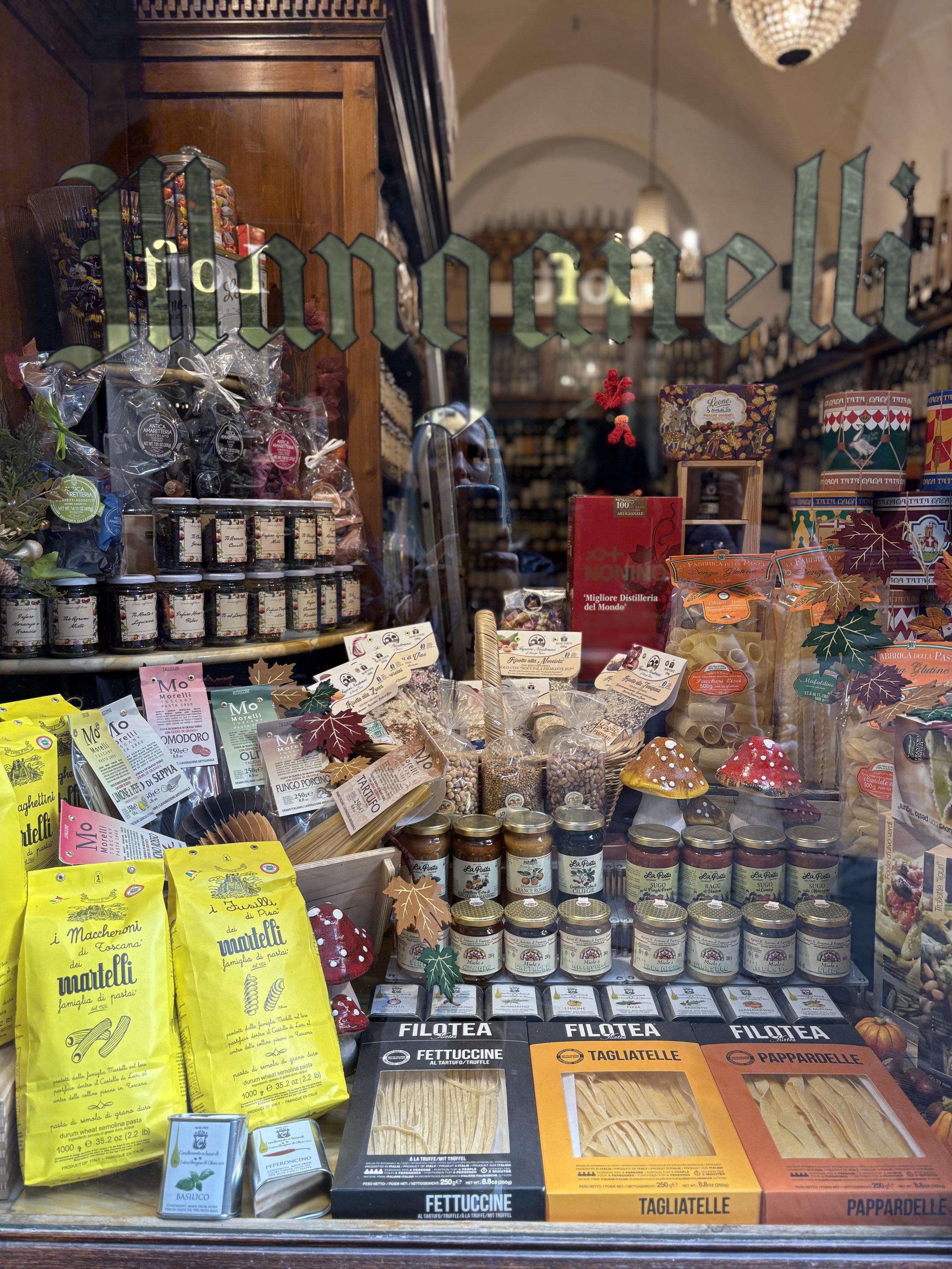 A bakery window display with various pasta, jars, and packaged food items, decorated with festive leaves and mushrooms, with shelves of wine bottles in the background.