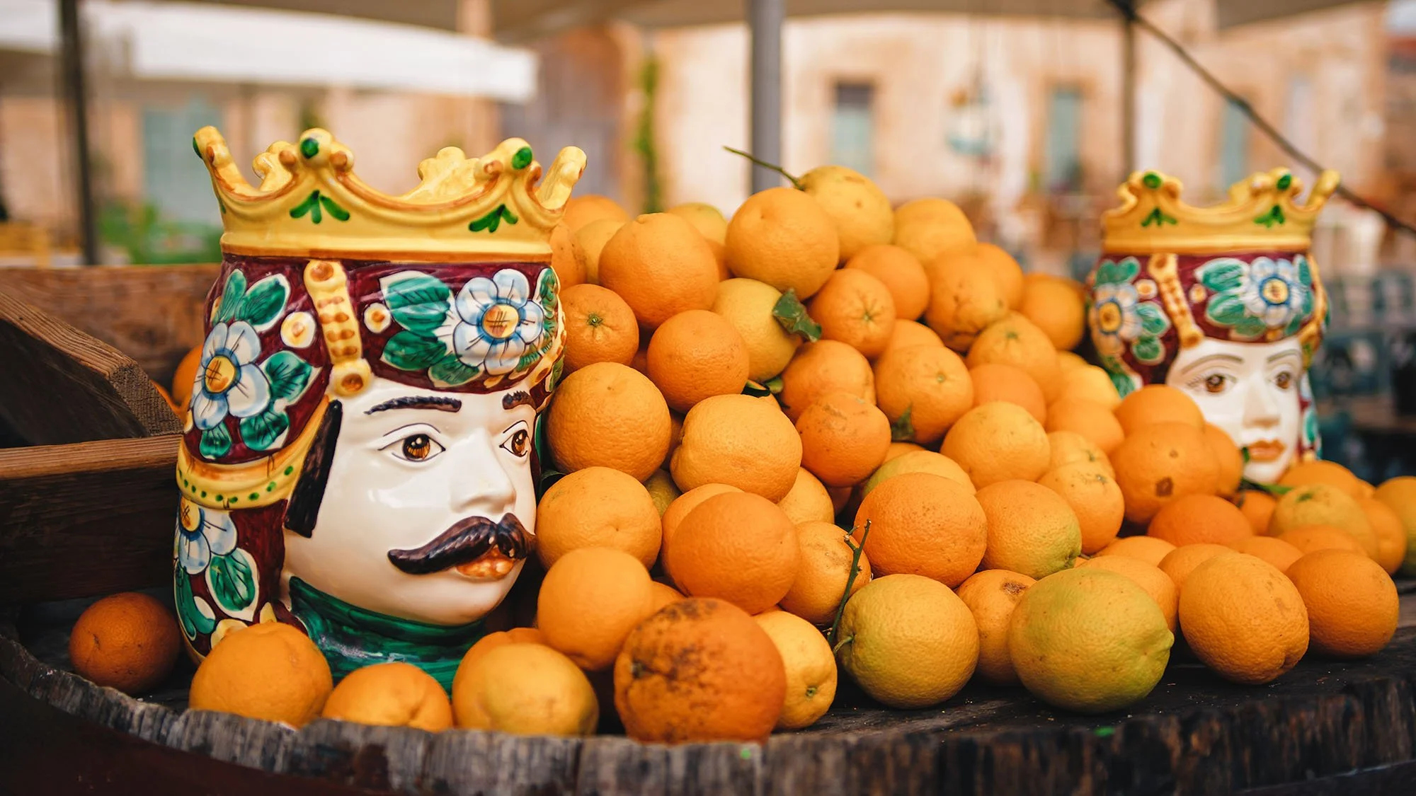 Colorful ceramic masks of a king and queen surrounded by a large pile of oranges on a wooden surface, likely at an outdoor market.