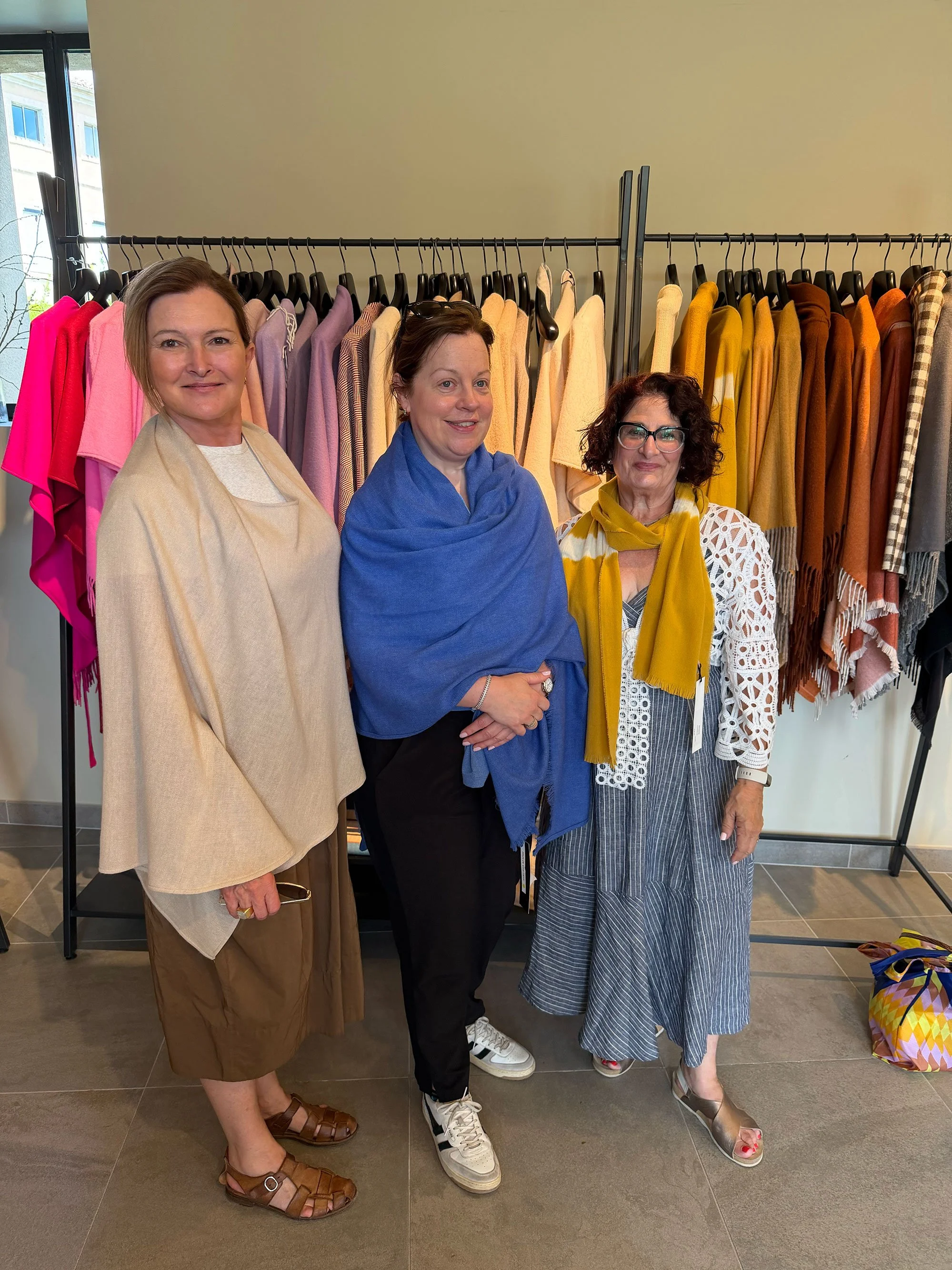 Three women standing together in front of a clothing rack with colorful sweaters, indoors with a window showing a building outside.