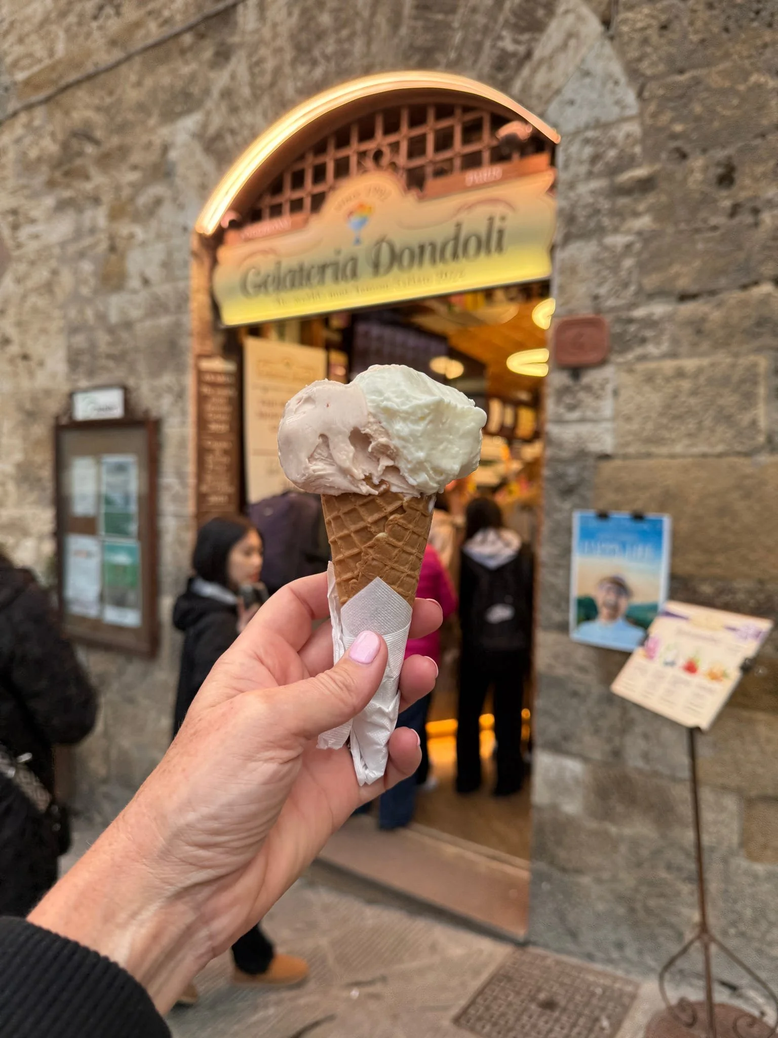 Hand holding a waffle cone with two scoops of ice cream in front of an ice cream shop entrance.
