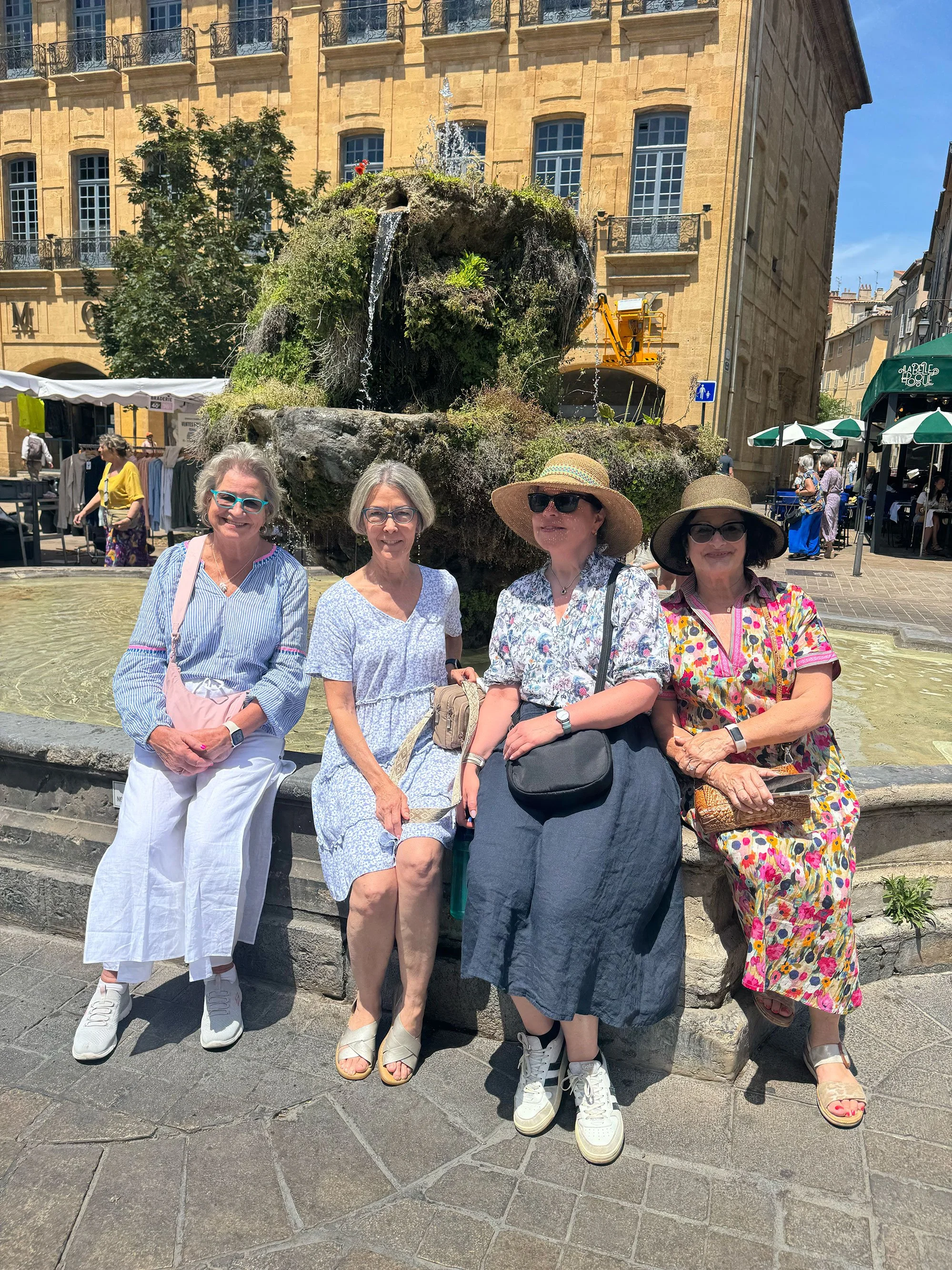 Four women sitting and standing together by a fountain in a busy city square. They are wearing hats, sunglasses, and colorful clothing, with a historic building and marketplace stalls in the background.