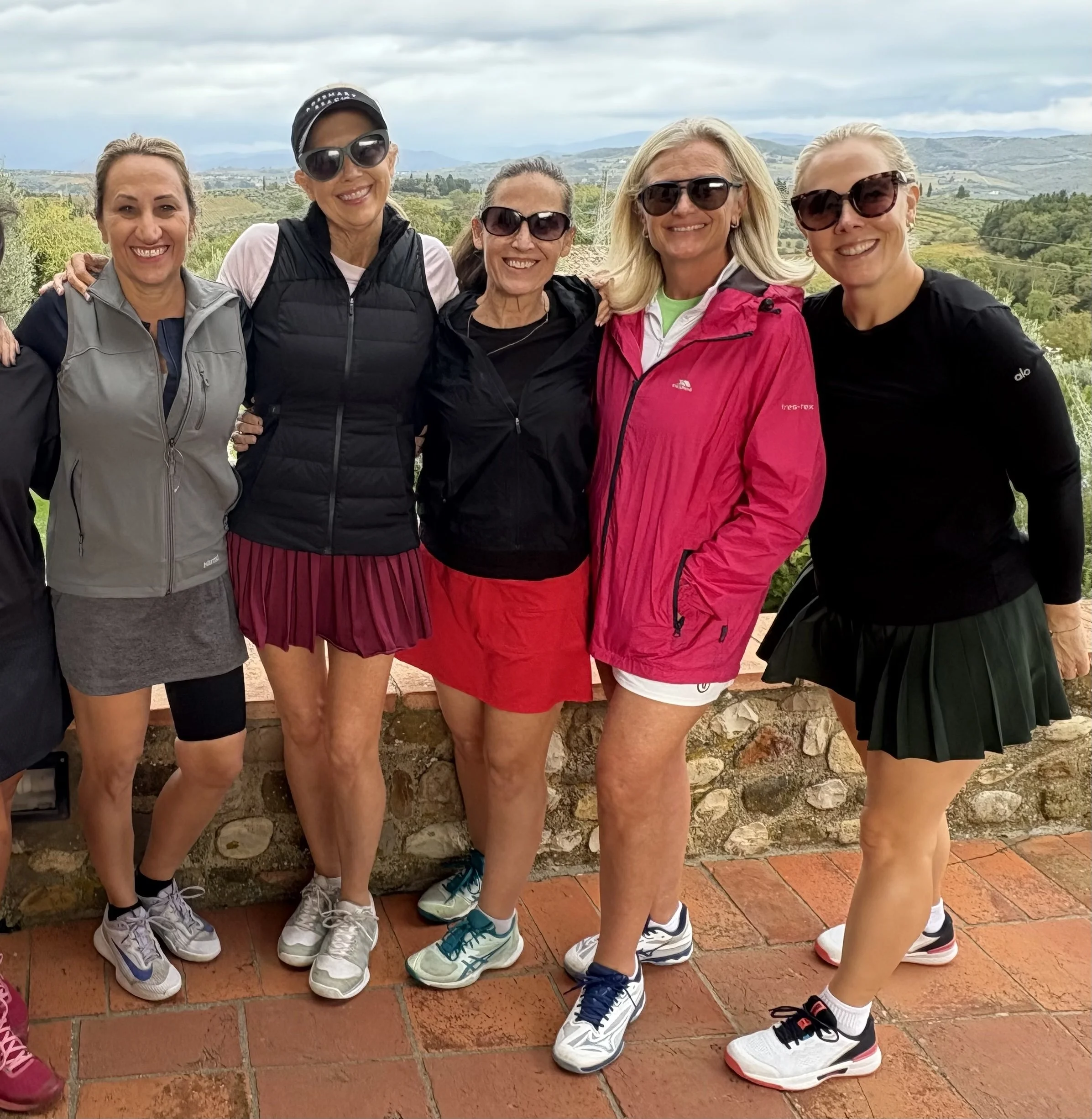 Group of five women standing outdoors on a balcony with scenic rolling hills and cloudy sky in background, dressed in athletic or outdoor gear, smiling and posing for the camera.