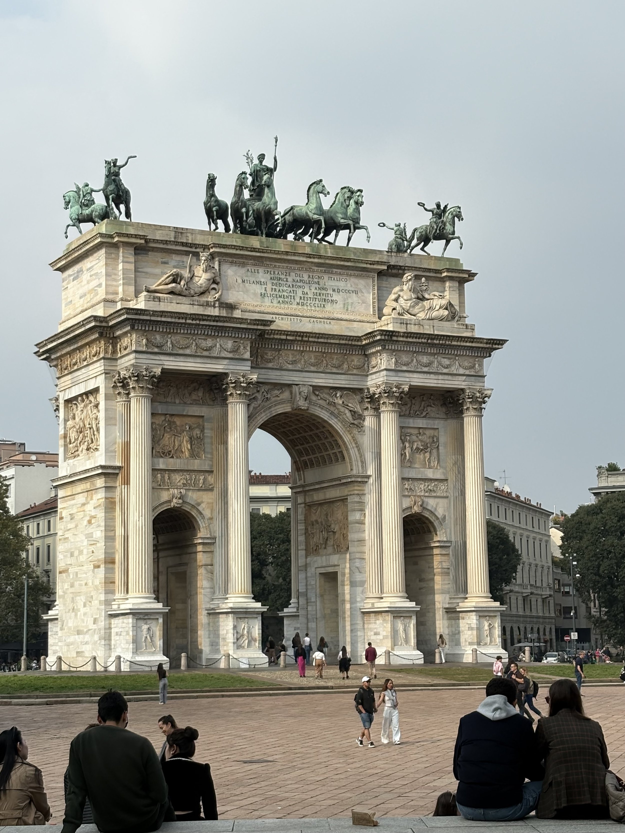 People sitting and walking around the Arco della Pace, a large white arch with Corinthian columns and a bronze chariot sculpture on top, in Milan, Italy.