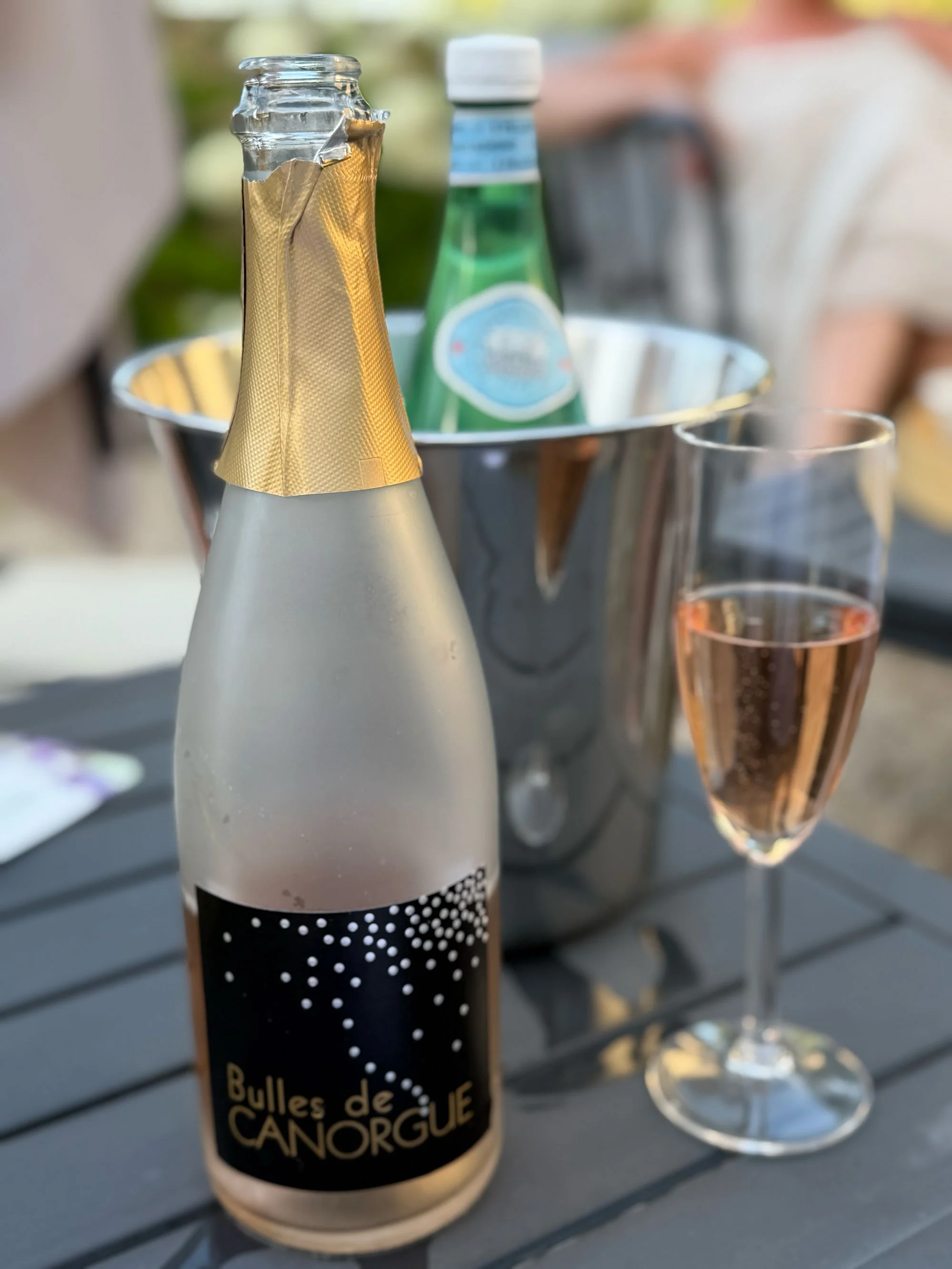 A bottle of champagne with a gold foil capsule and a black label with white dots, a glass of pinkish bubbly, and a green bottled beverage in an ice bucket on a gray outdoor table.