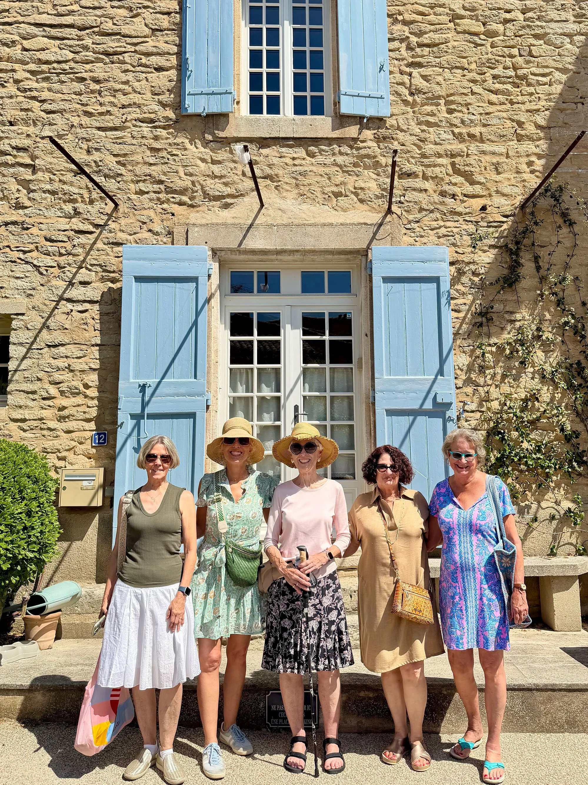 Group of six women, smiling, standing in front of a stone building with light blue shutters and window frames, some women wearing hats, sunglasses, and casual summer clothing, on a sunny day.