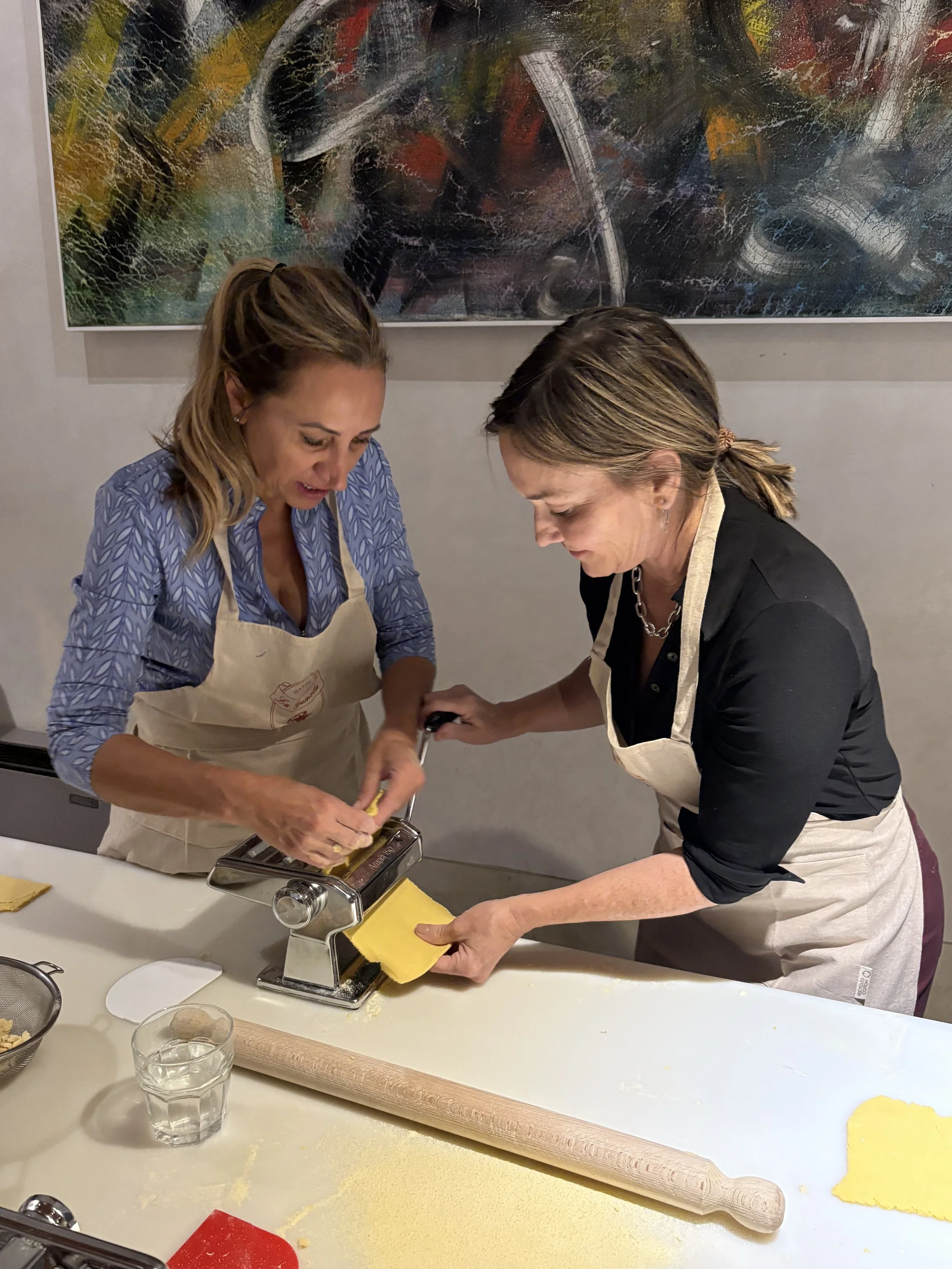 Two women in aprons making pasta with a pasta machine on a white table, with a rolling pin and glass of water nearby.