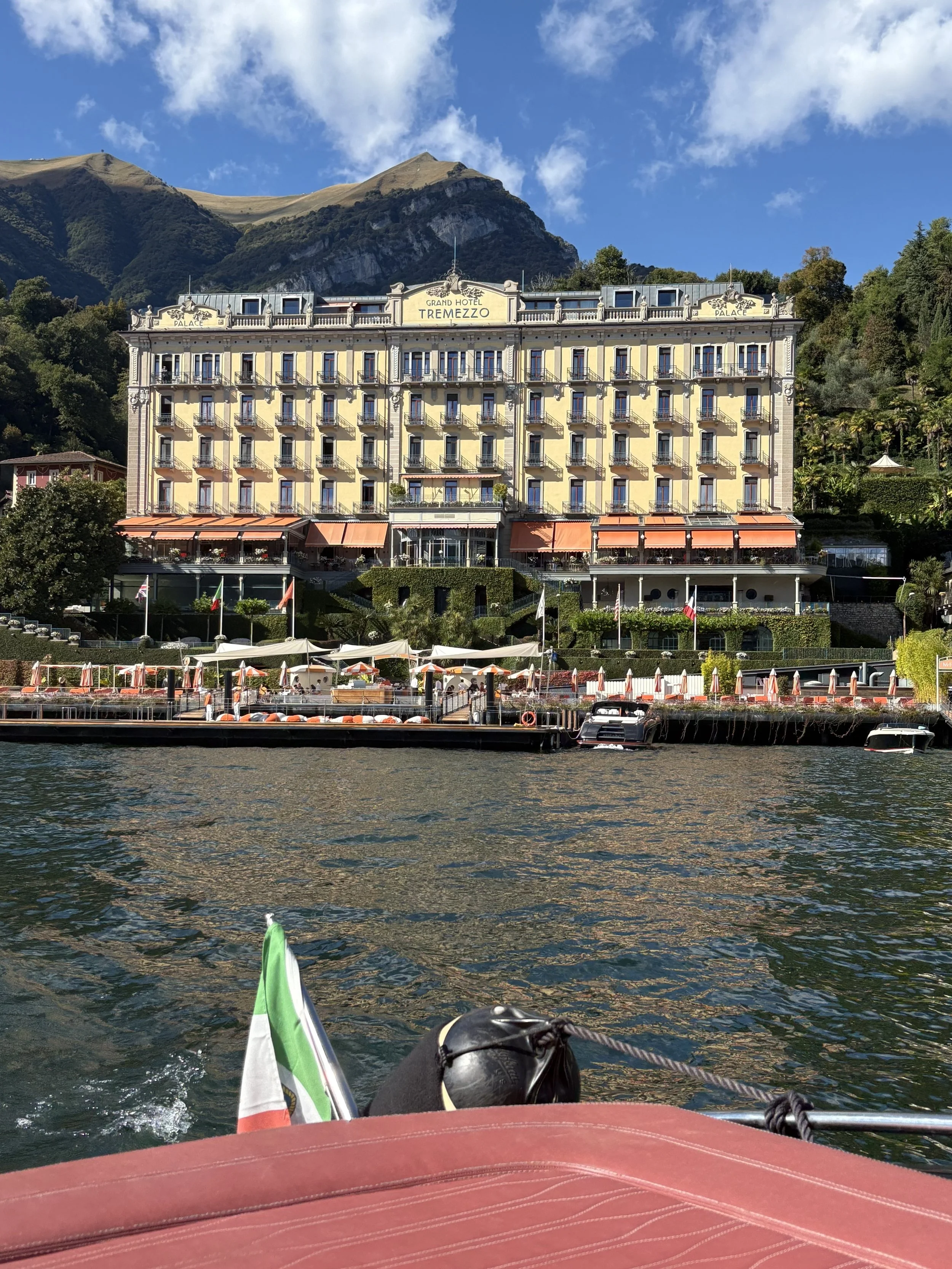 View of Lake Como with a boat in the foreground, showing the Grand Hotel Tremezzo on the shoreline with mountain backdrop and blue sky.