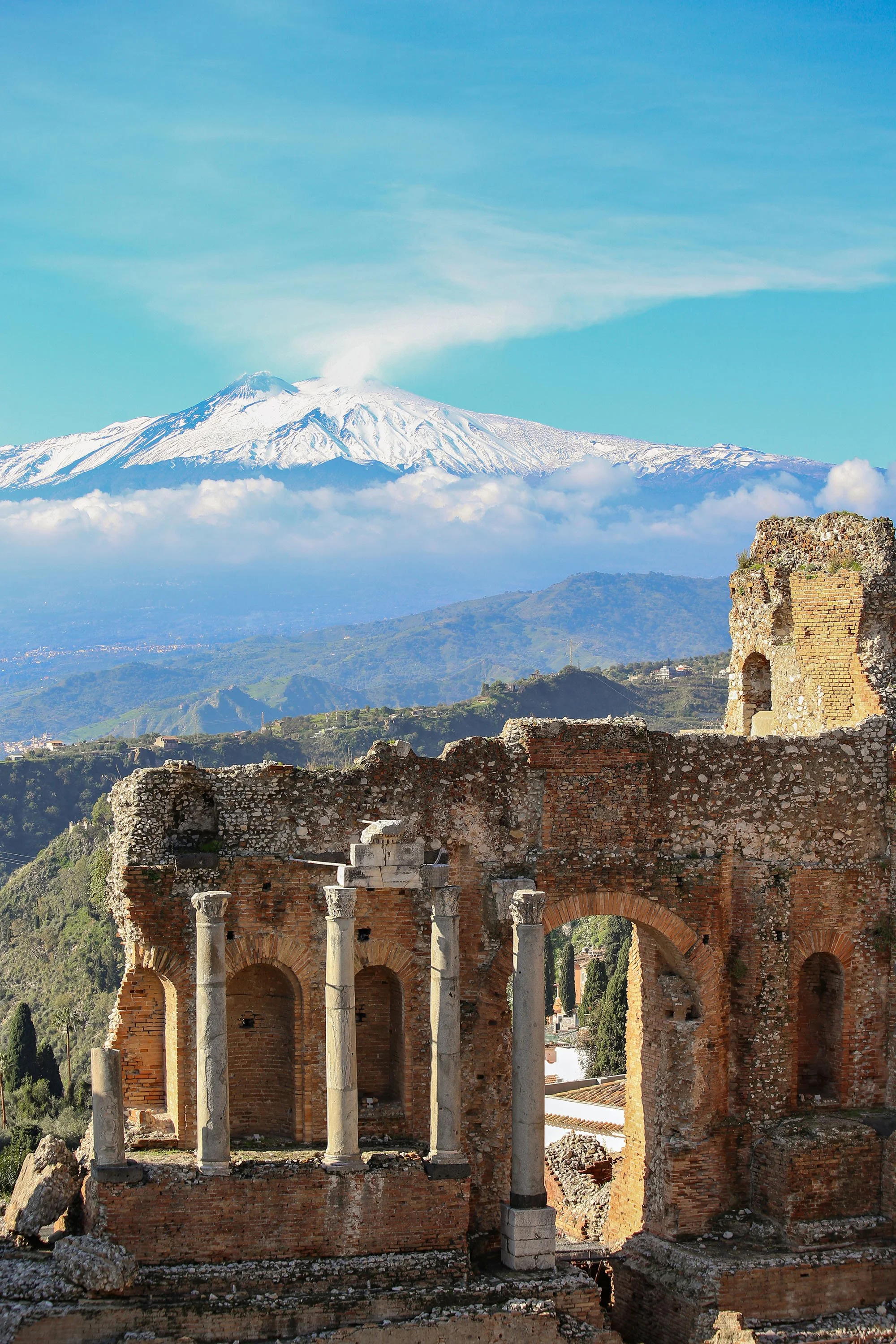 Ancient ruins with three tall white marble columns and brick arches, with snow-capped Mount Etna volcano erupting in the background under a blue sky.