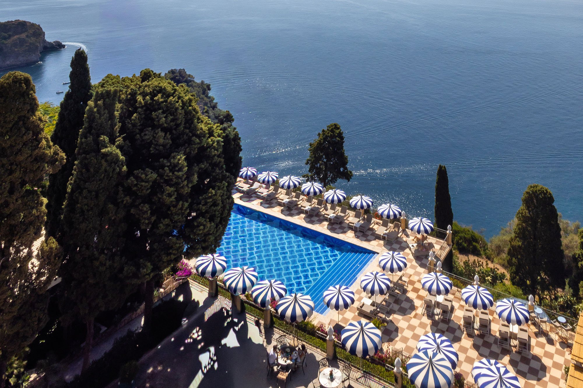 A swimming pool area with striped umbrellas and lounge chairs, overlooking a body of water with cliffs and greenery in the distance.