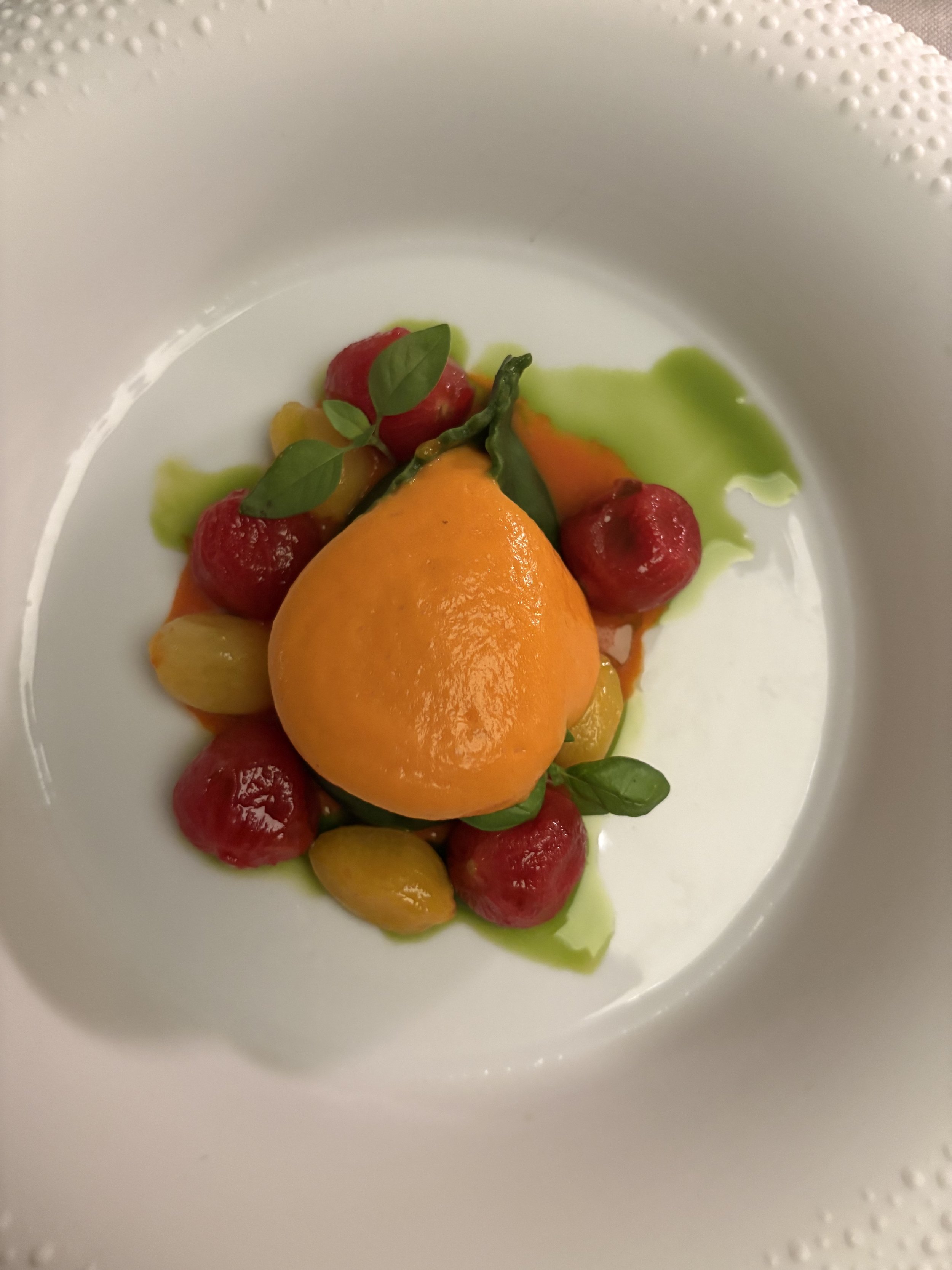 A plate of colorful dessert featuring a large peach in the center, surrounded by red and yellow fruit balls, green sauce, and garnished with small fresh green leaves.