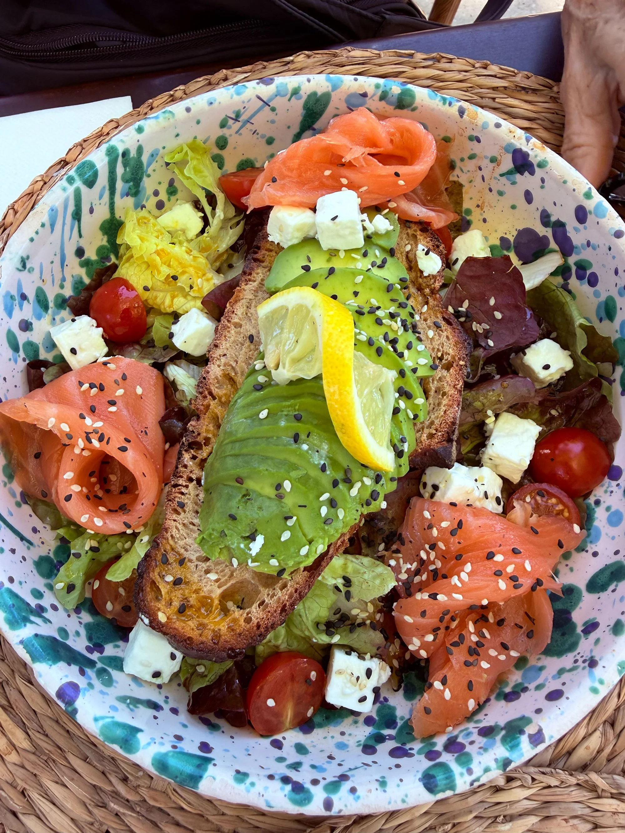 A colorful plate of open-faced smoked salmon and avocado toast with lemon, feta cheese, and sesame seeds, accompanied by a side salad with cherry tomatoes, cheese, and lettuce.