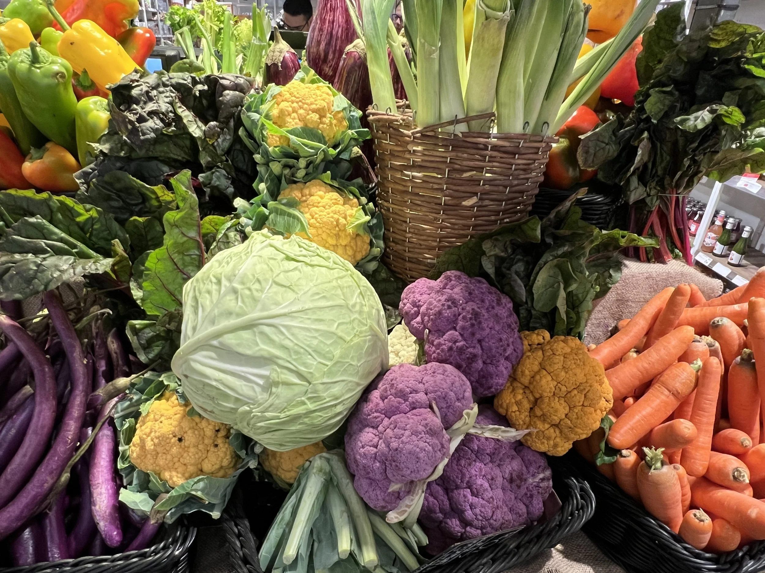 A display of fresh vegetables at a market, including purple cauliflower, yellow cauliflower, green cabbage, carrots, eggplant, leeks, and leafy greens.