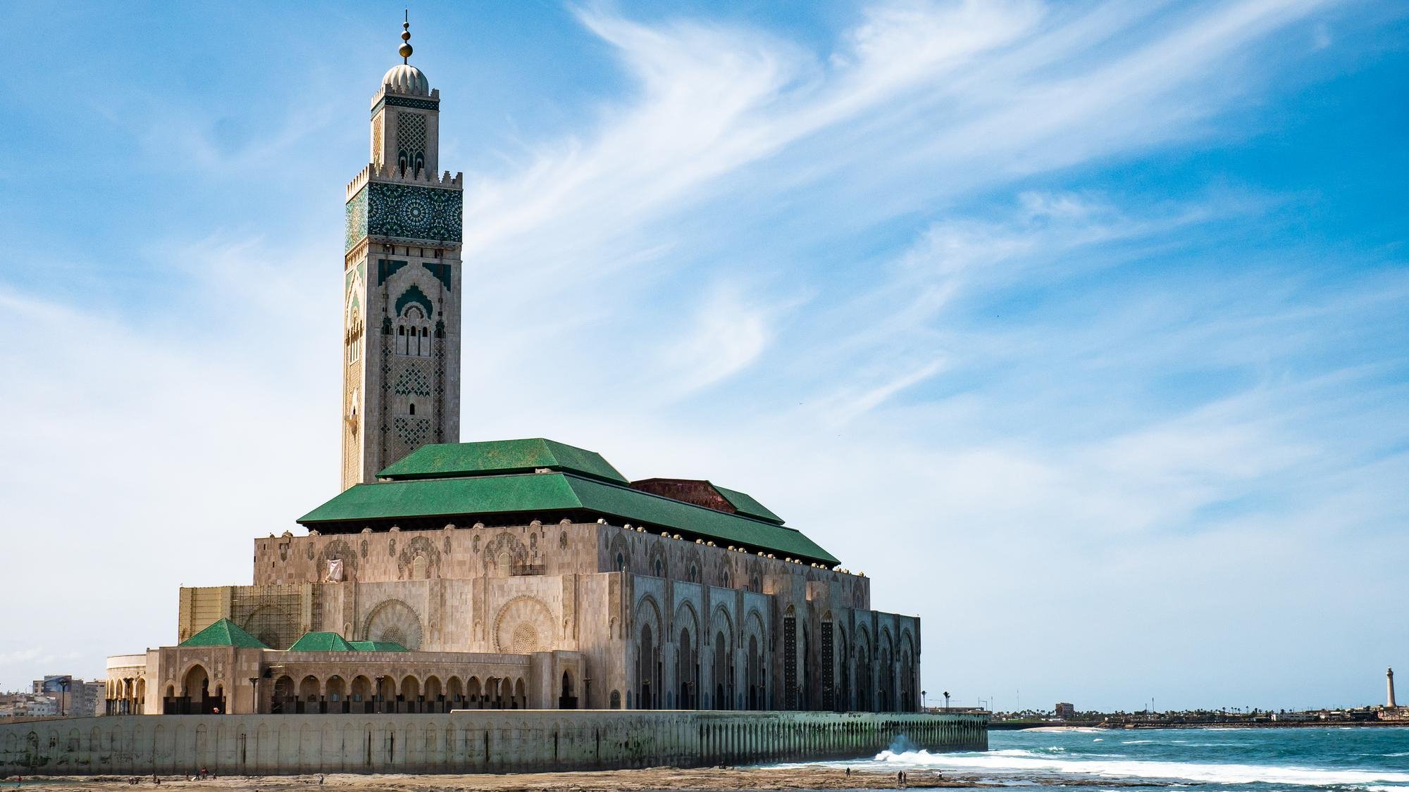 The Hassan II Mosque in Casablanca, Morocco, with its minaret, green rooftops, and ocean waves, under a blue sky with wispy clouds.