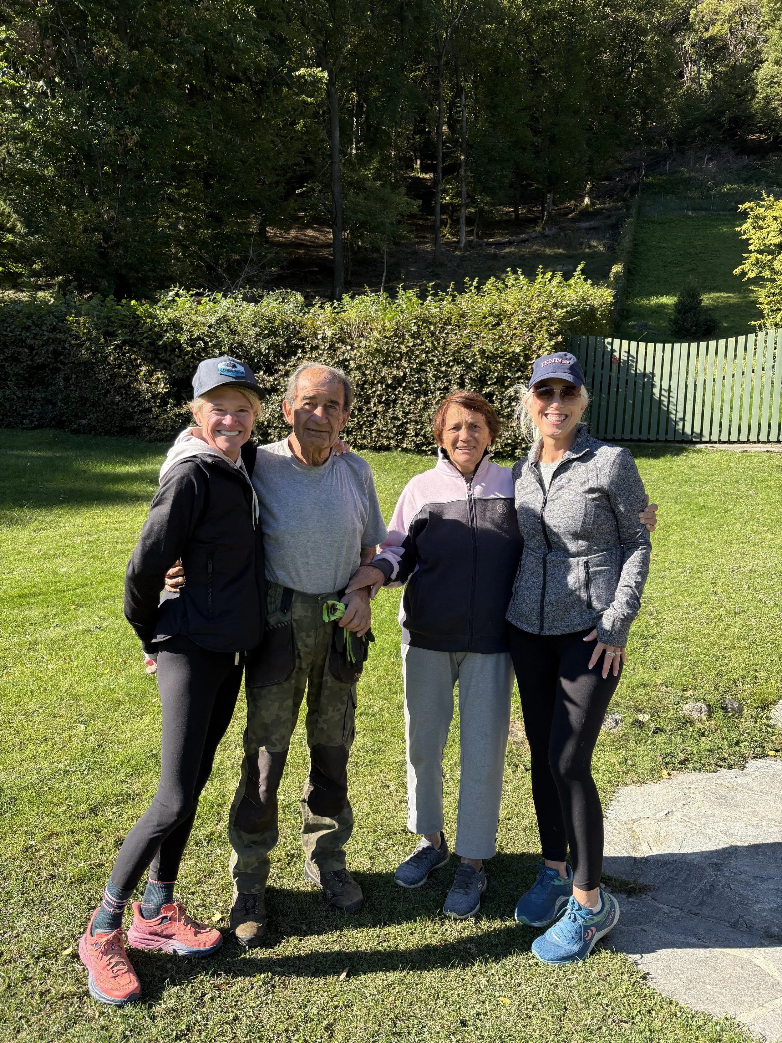 Four women and one man standing together outside in a grassy area with trees and a green fence in the background, smiling at the camera.