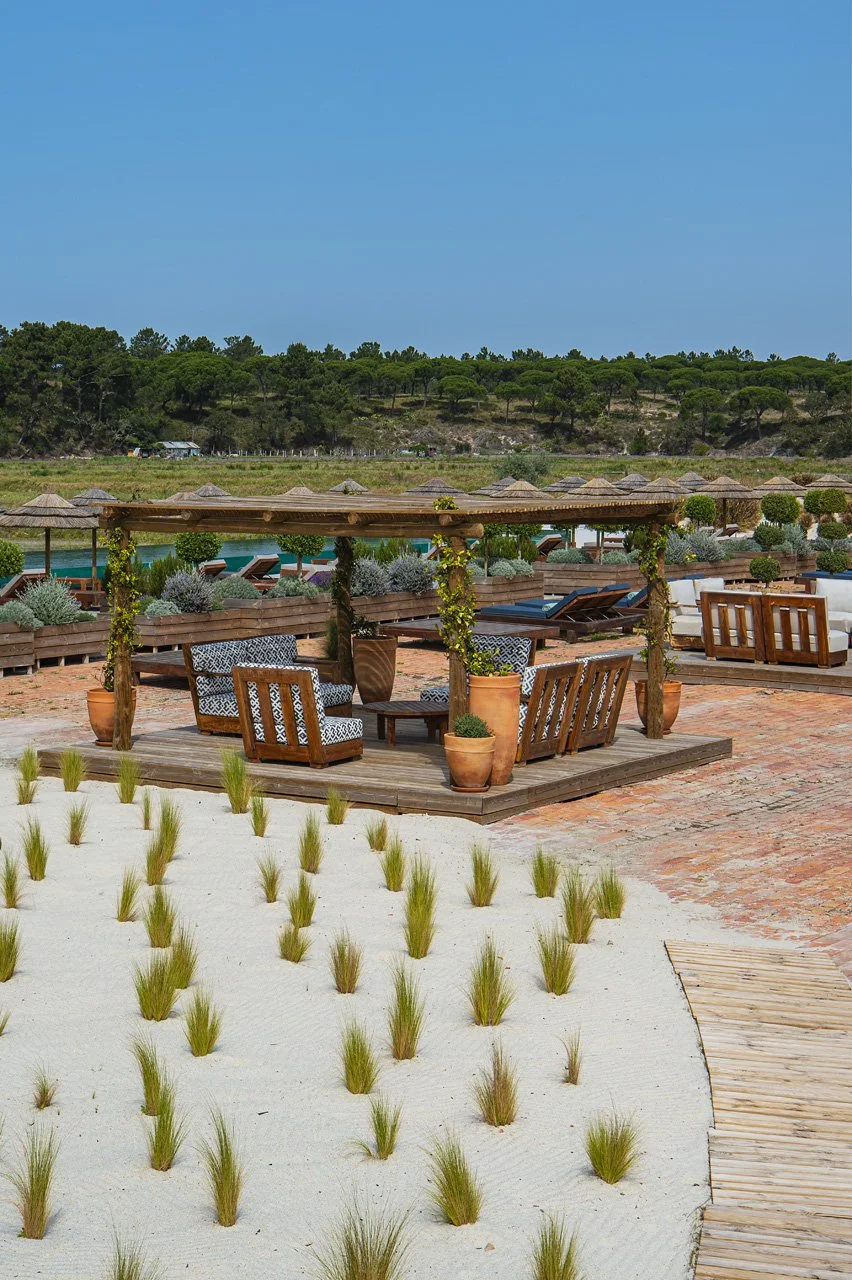 Patio area with wooden seating, potted plants, and umbrellas on a sunny day, with a field and trees in the background.