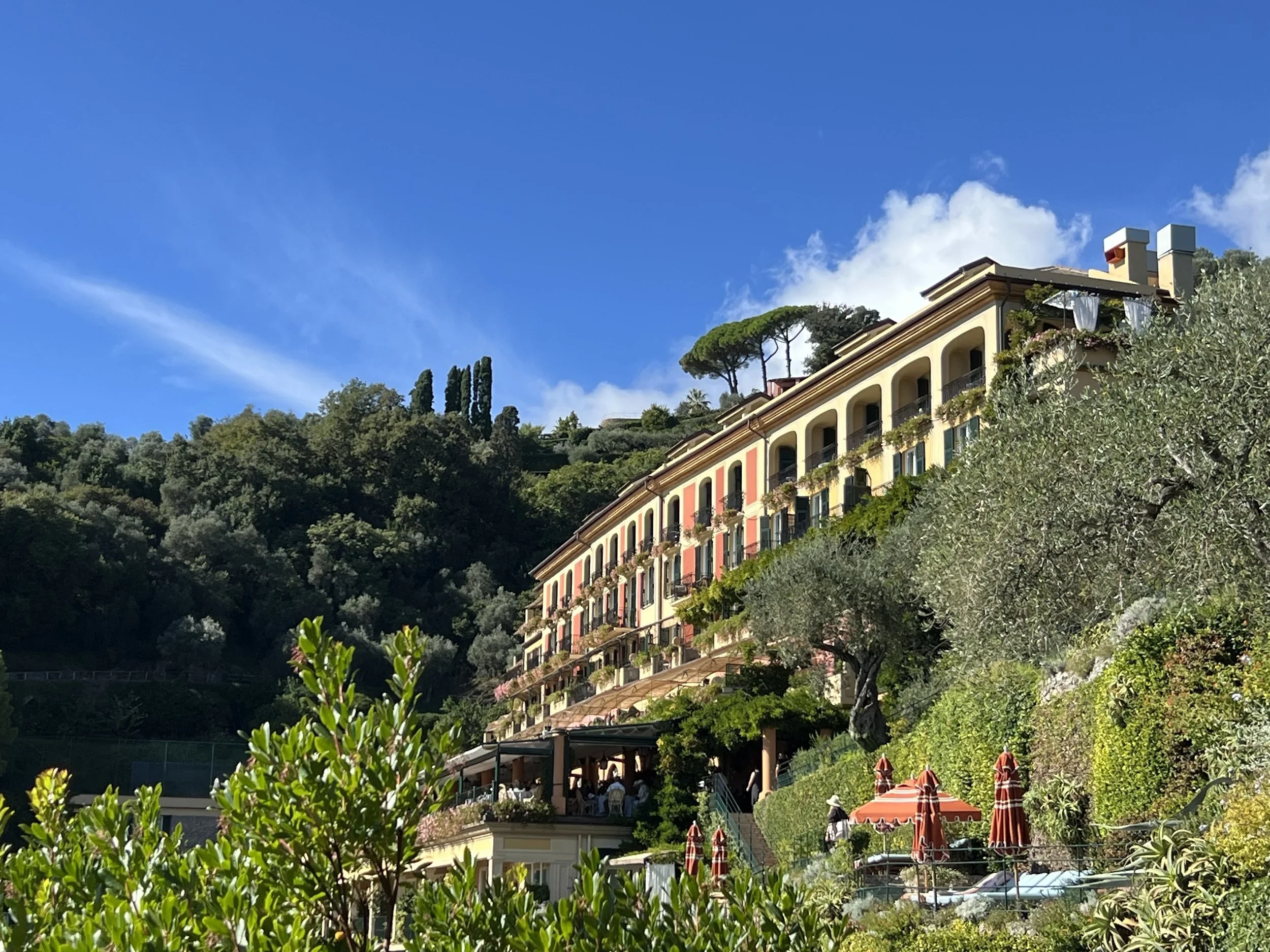 Colorful building on a hillside with greenery, trees, and shrubs, under a blue sky with some clouds.