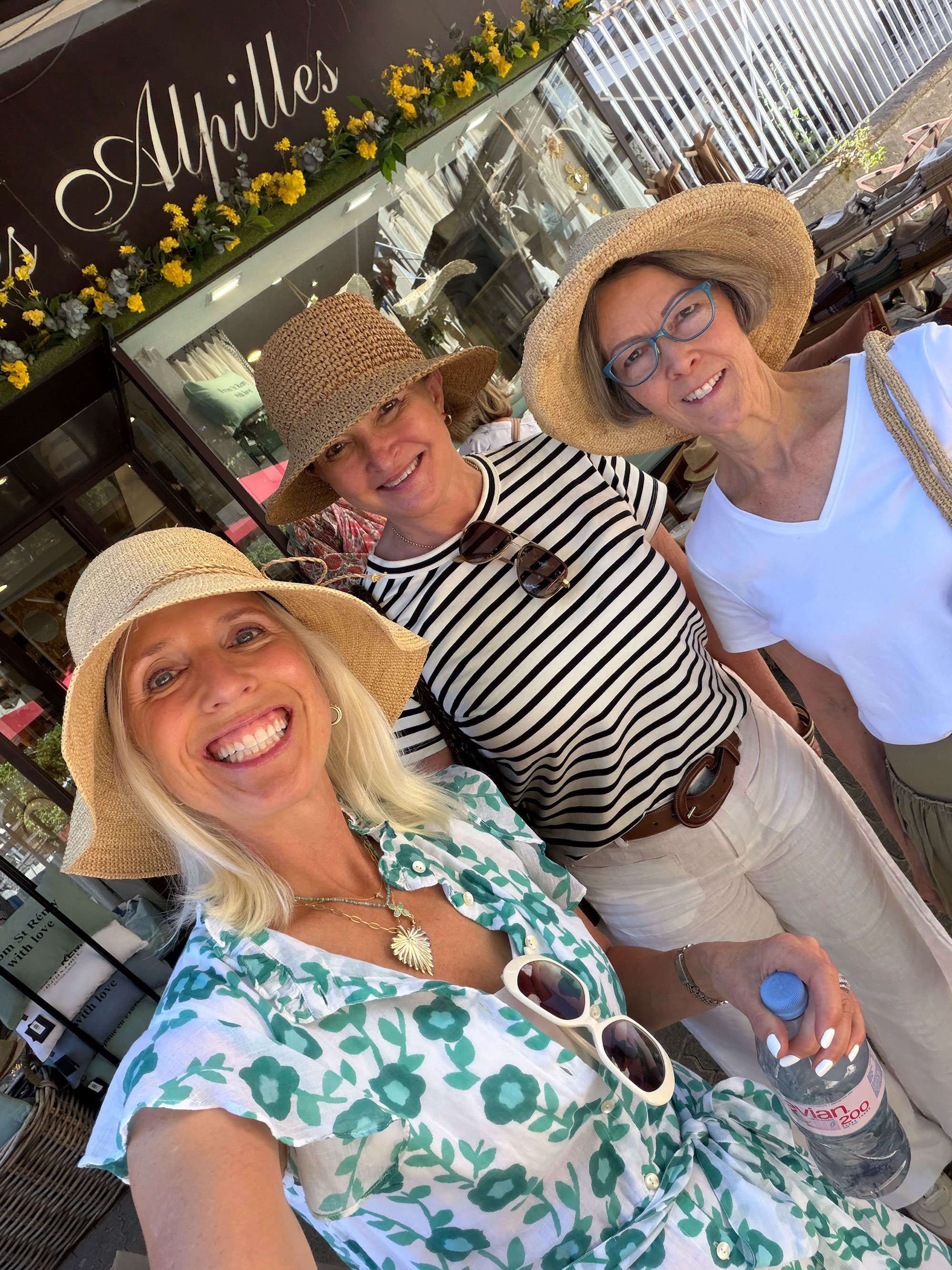 Three women wearing large straw hats and smiling for a selfie outdoors. They are dressed in light, summery clothing and one is holding a bottle of water. The background shows a storefront with flowers and a metal fence.
