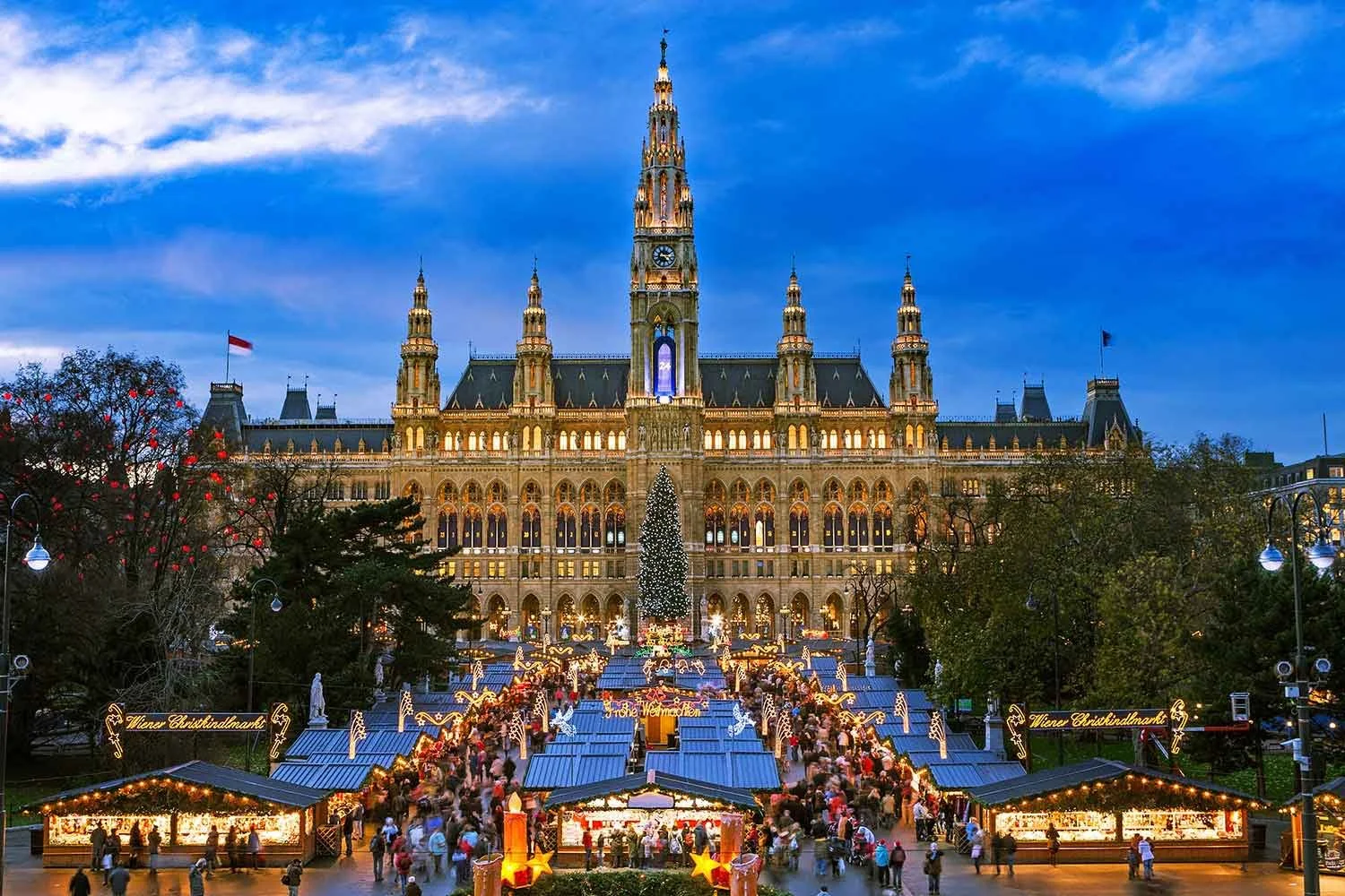Christmas market scene in front of a historic building with a tall illuminated clock tower, decorated Christmas tree, festive lights, and crowds of people shopping at market stalls.